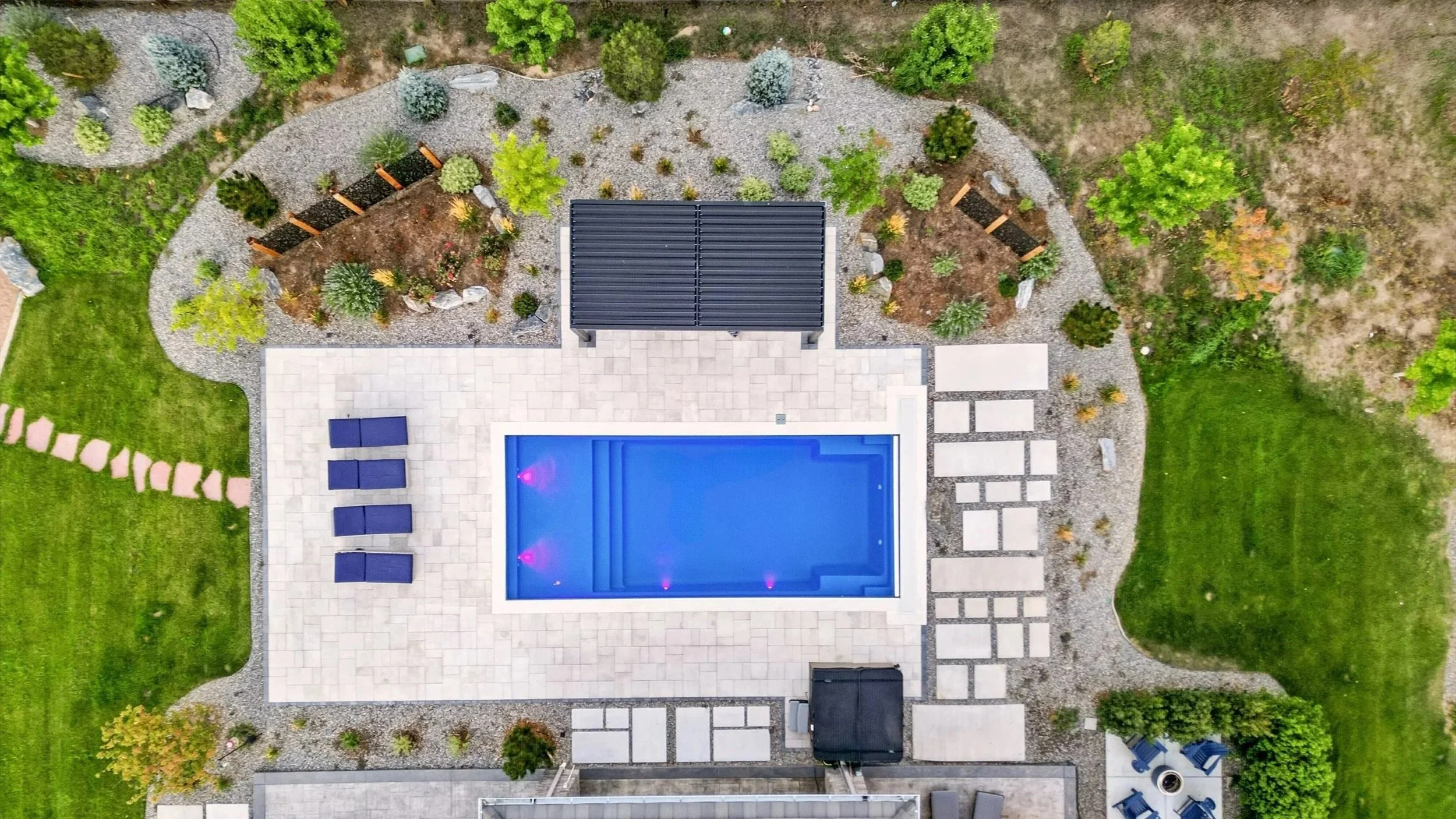 An aerial view of a backyard with a swimming pool, patio chairs, landscaped garden beds, and surrounding grass and trees.