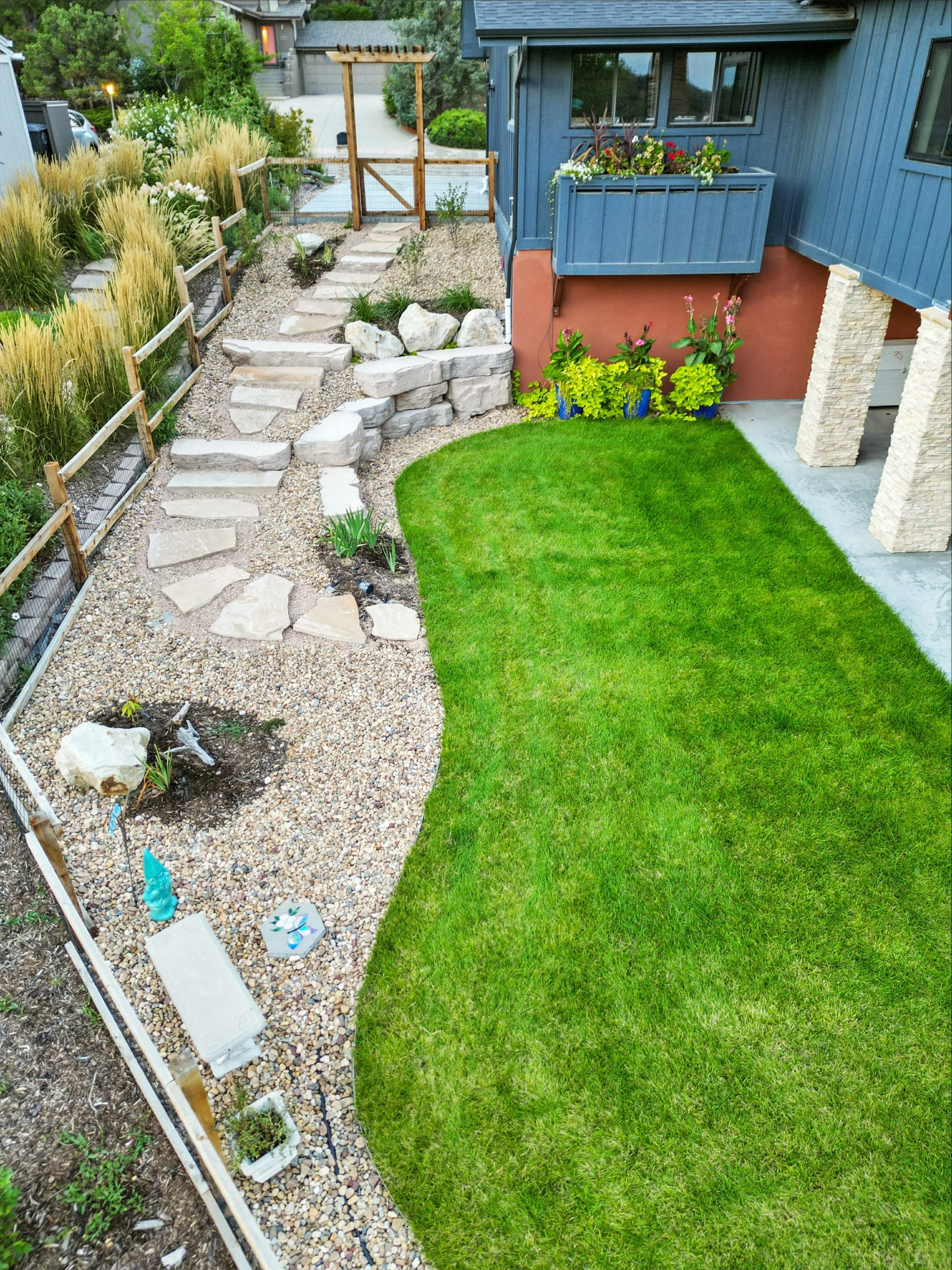 A landscaped backyard with a stone pathway, a small grassy lawn, and various plants and flowers. There is a blue house wall and a balcony with flower pots on the right, with a wooden gate and a driveway in the background.