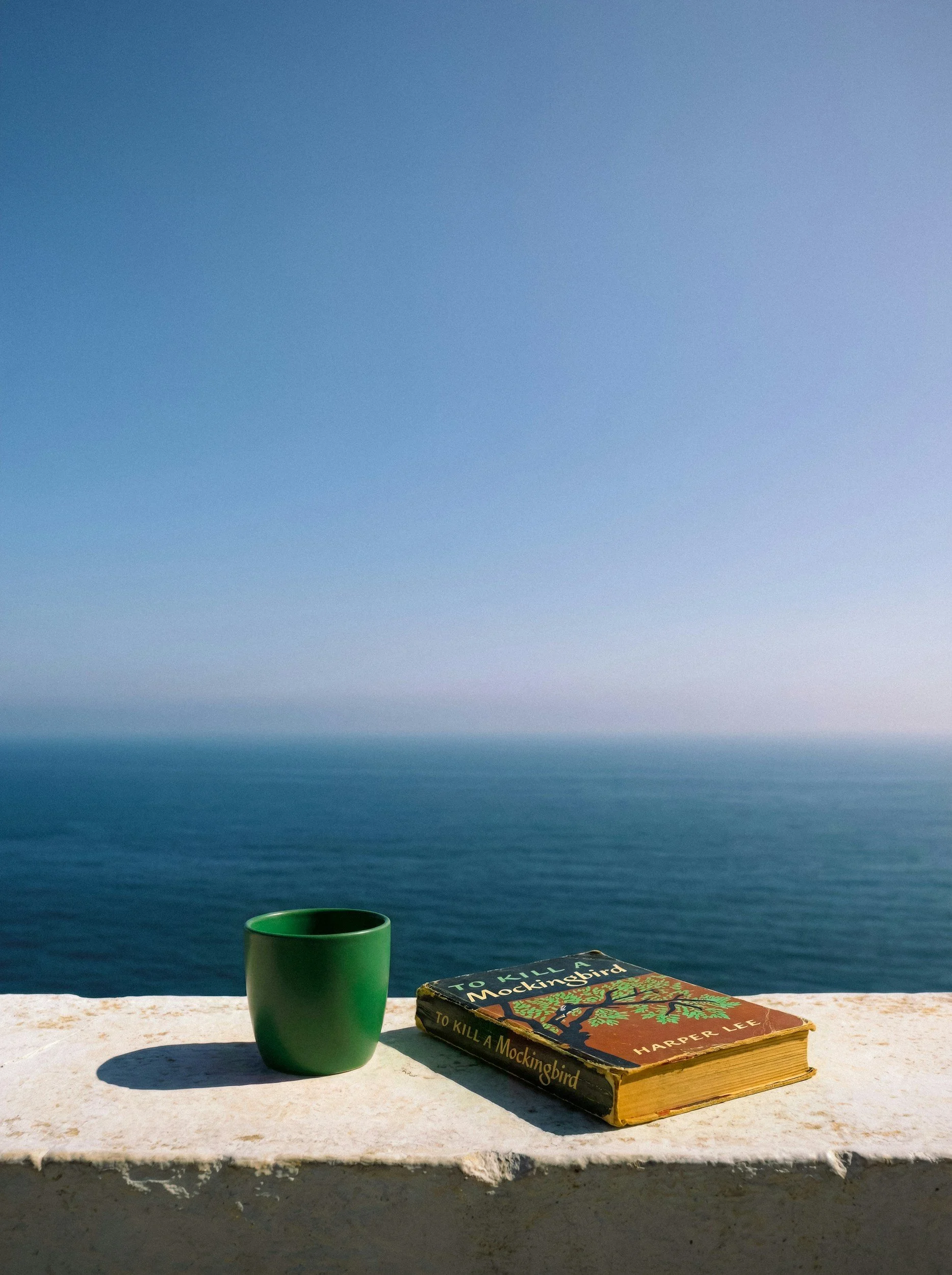 A green mug and a worn copy of 'To Kill a Mockingbird' by Harper Lee on a windowsill overlooking the ocean with a gradient sky.