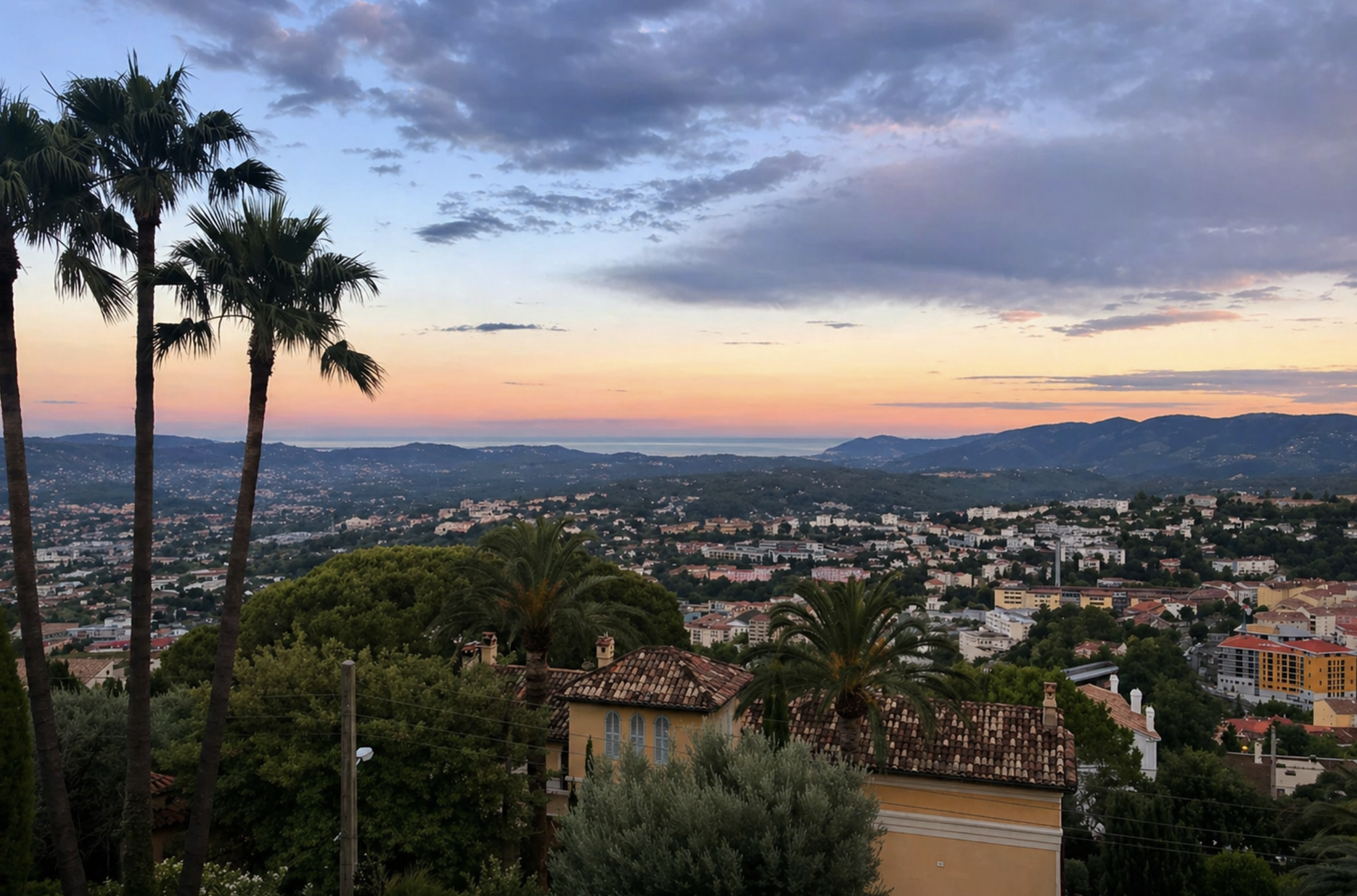 View of a cityscape at sunset with a backdrop of hills and a cloudy sky, framed by tall palm trees and lush greenery in the foreground.