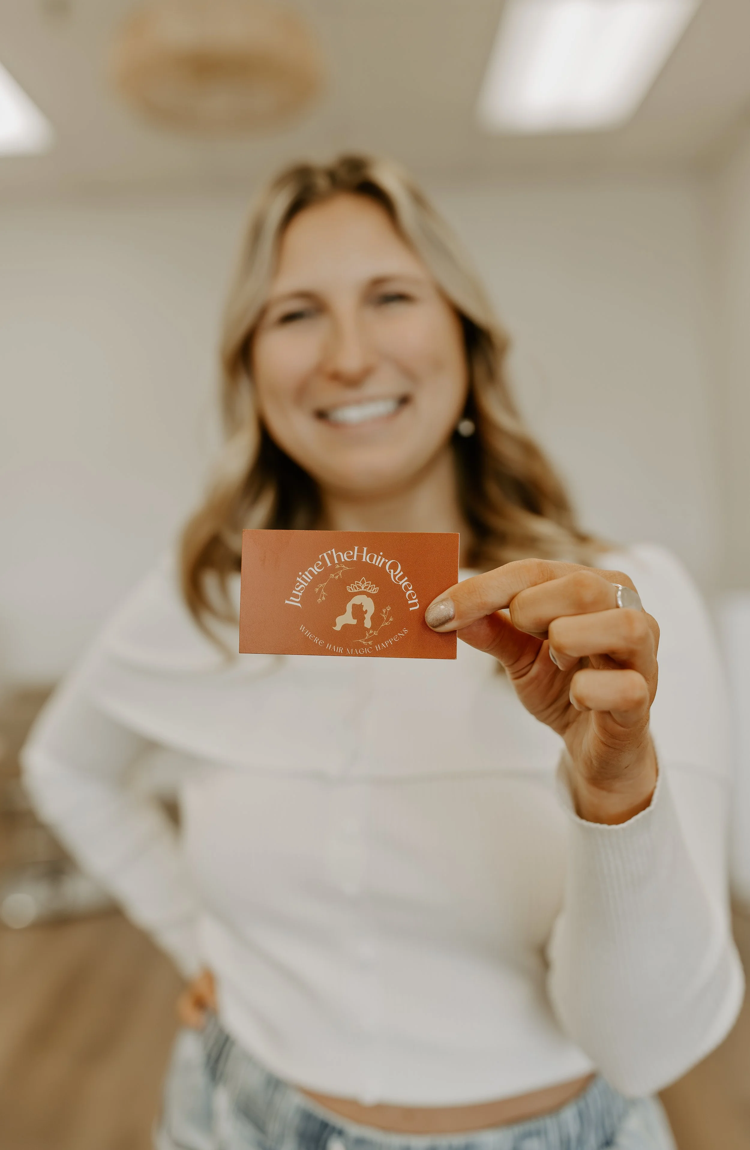 Woman in white shirt holding a business card that reads 'Justine The Hair Queen' with a silhouette of a woman with a crown.