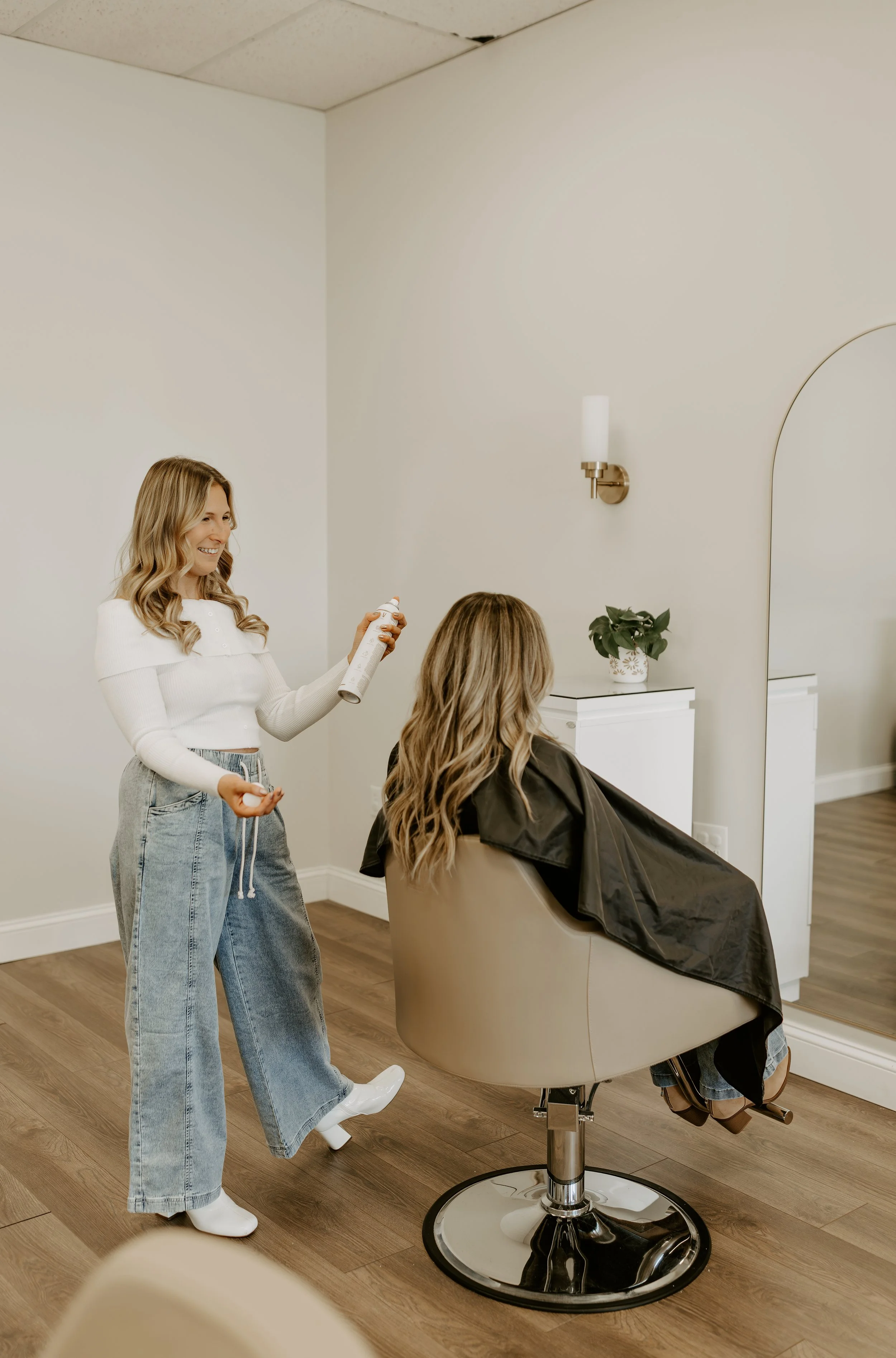 A woman sitting in a salon chair getting her hair styled, with a hairdresser standing nearby holding a can of hair spray and smiling.