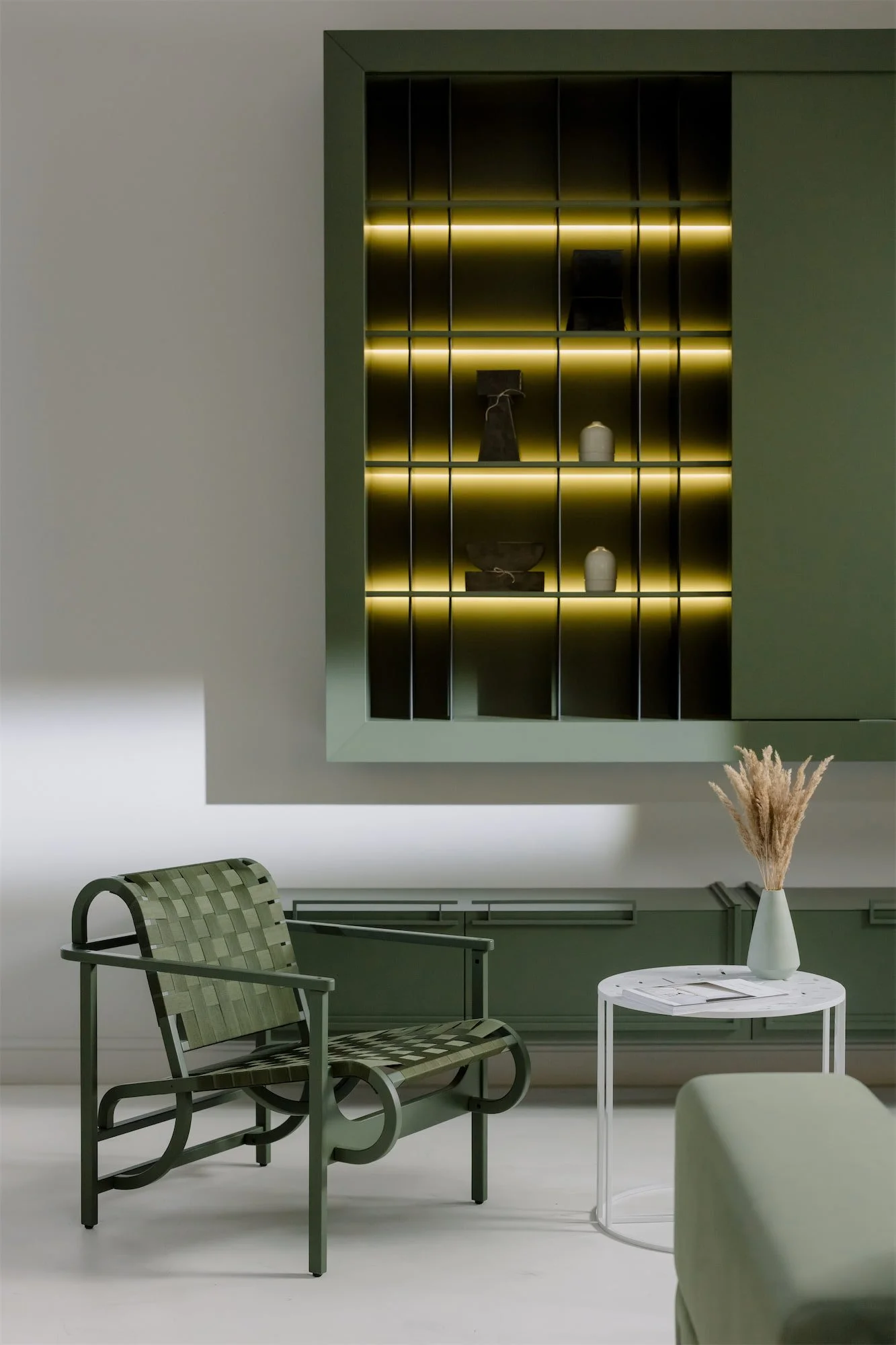 A green wall-mounted display cabinet with decorative items inside, including vases and a bowl, illuminated by warm lighting. Below, there's a green metal-framed chair with a woven seat and back, a small white round side table with a vase of dried pampas grass, and part of a white upholstered chair.