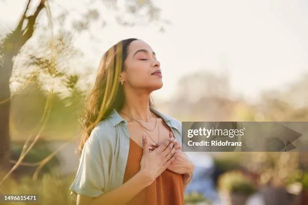 A young woman standing outdoors with eyes closed and hand on her chest, appearing to meditate or breathe deeply during sunset.