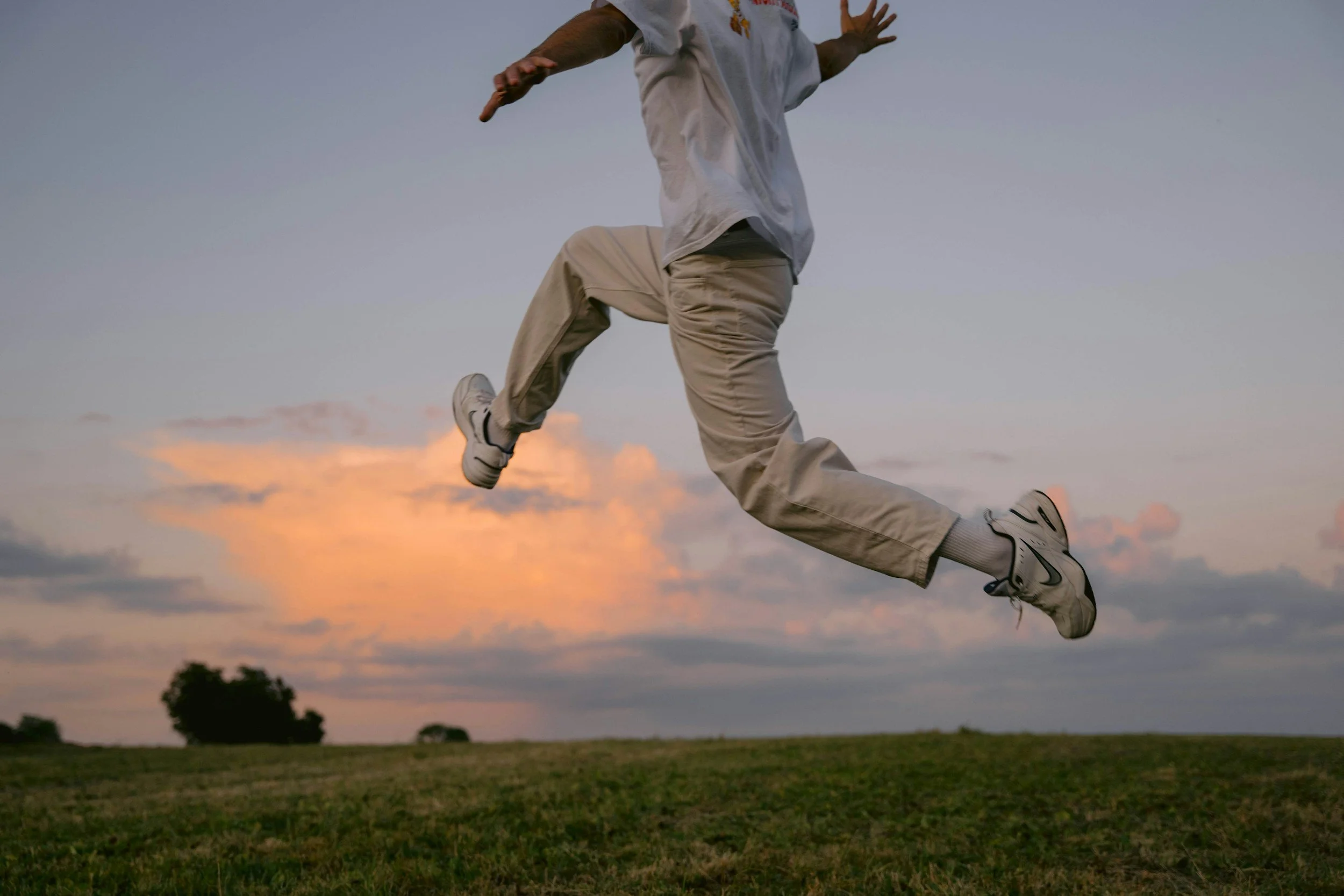 Person wearing white pants, a white shirt, and sneakers jumping in the air outdoors during sunset with a cloudy sky and a grassy field below.