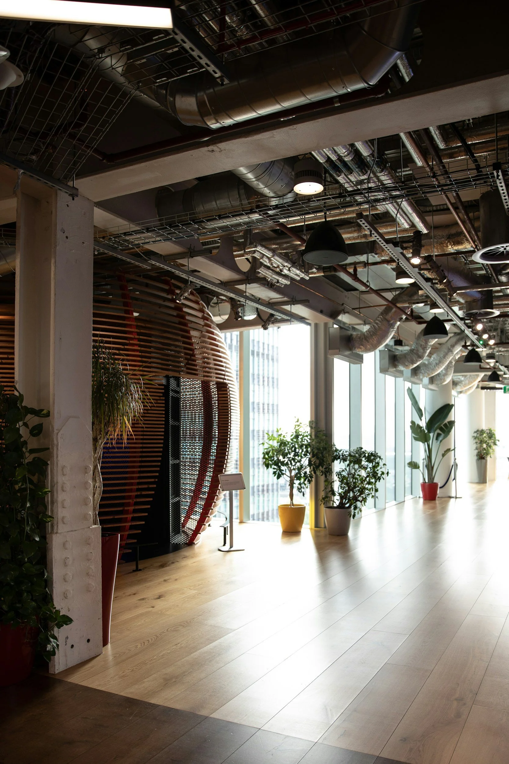 Interior of a modern office or building with wooden flooring, potted plants, large windows, and an exposed ceiling with industrial ductwork.