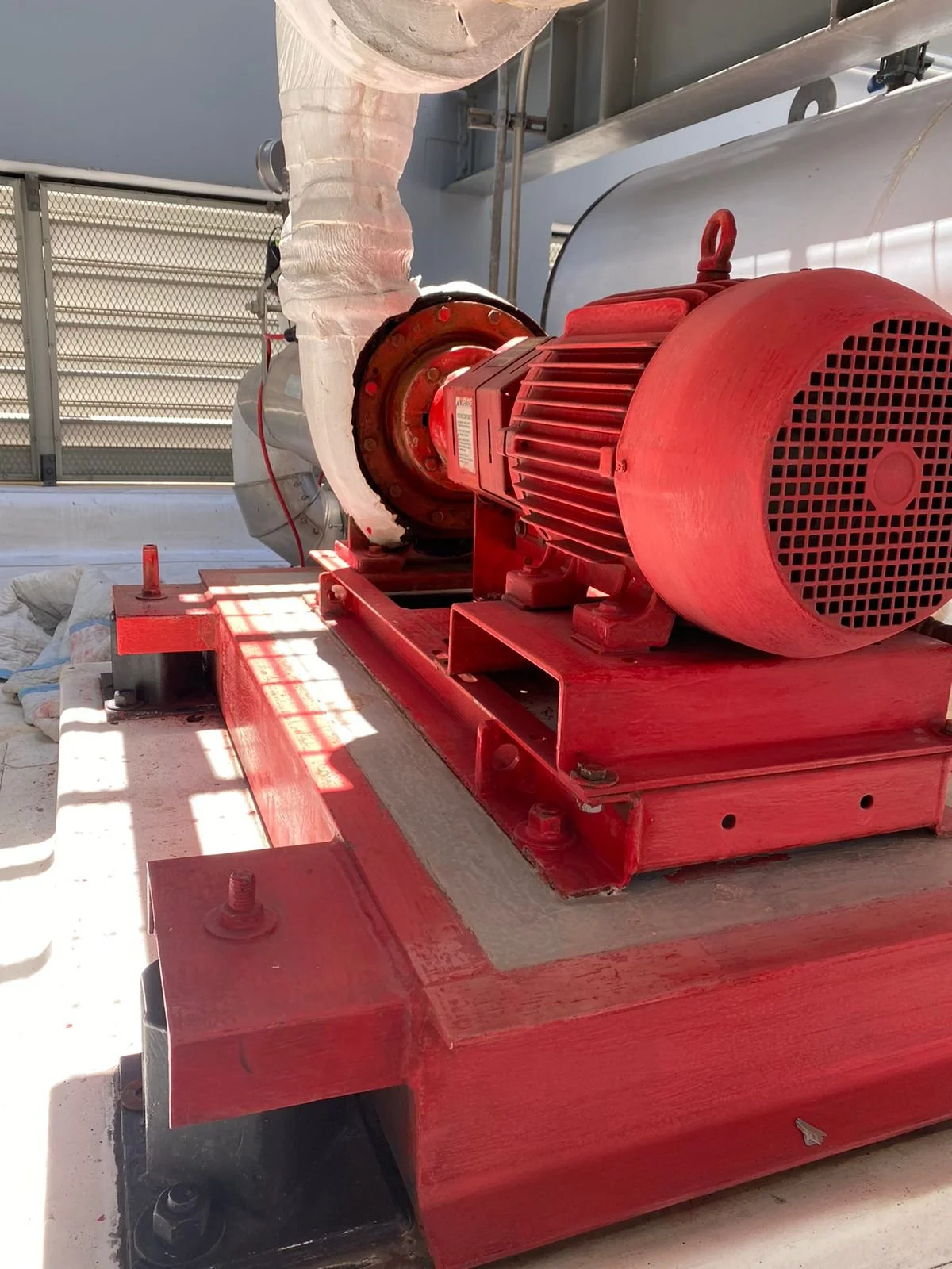 Bomba de agua roja instalada en una estructura metálica en un espacio abierto, con tubería gruesa blanca conectada a ella.