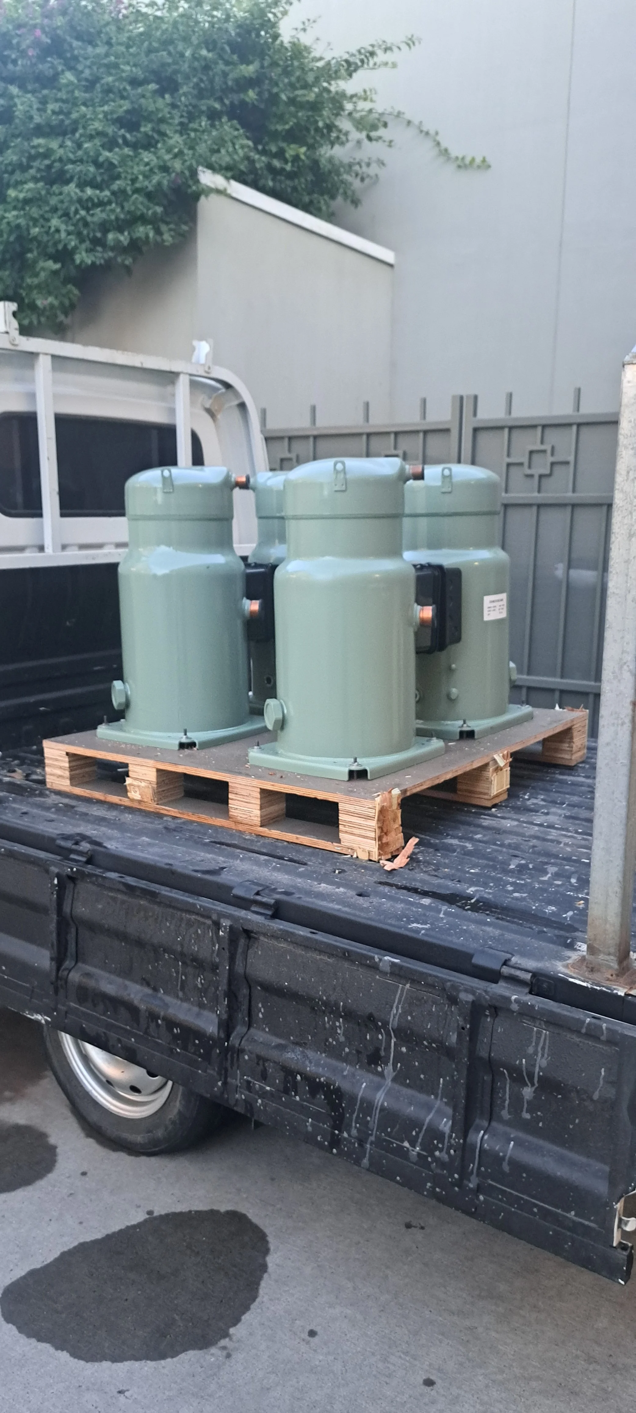 Cuatro compresores de refrigeración verdes en una plataforma de madera en la caja de un vehículo, con paredes y vegetación en el fondo.