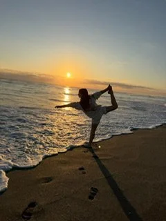 Person practicing yoga or a dance pose on the beach at sunset, with the ocean and sky in the background.