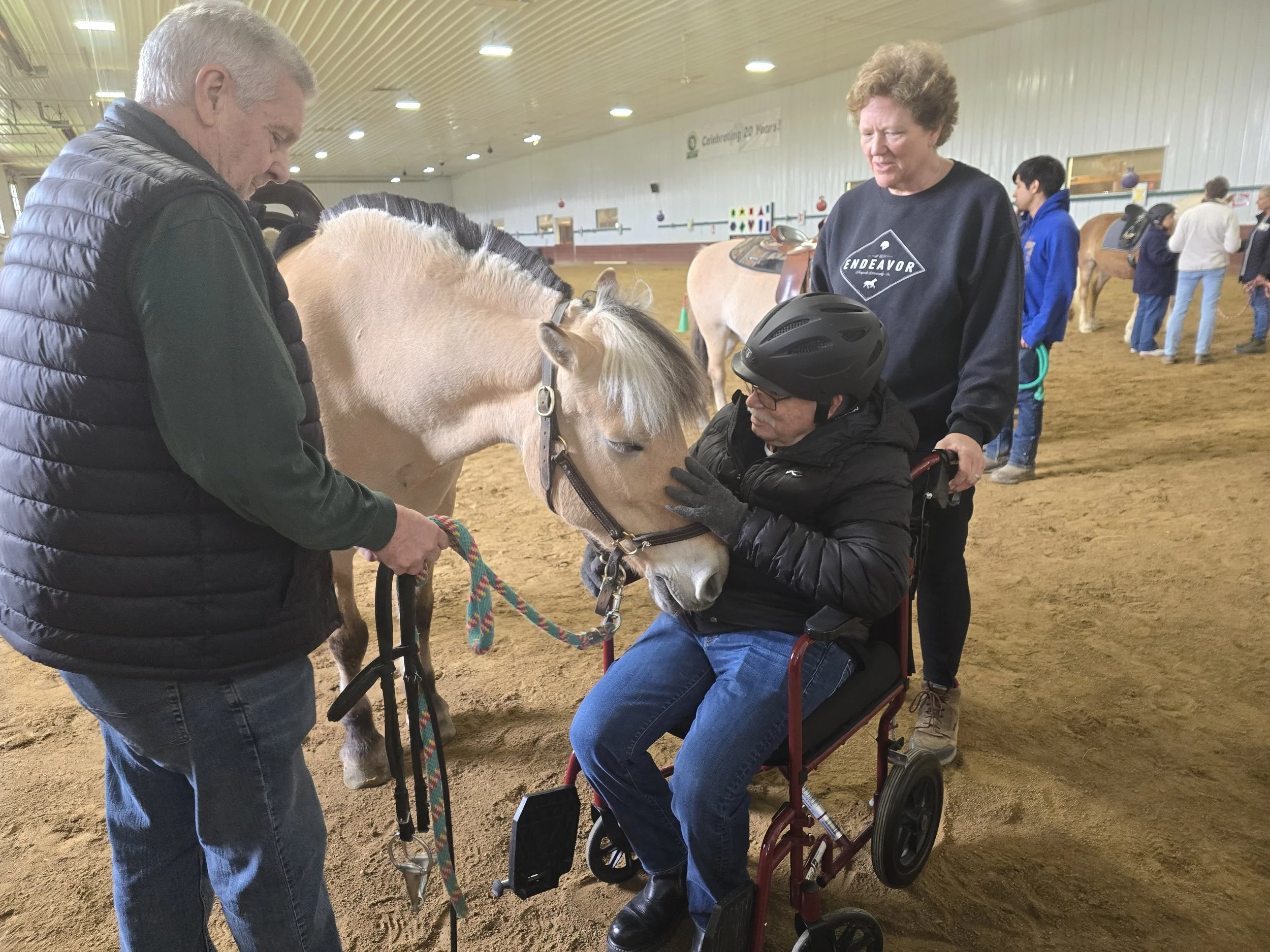 An indoor equestrian arena with four people gathered around a small white horse, one person is sitting in a wheelchair and wearing a helmet, while others are standing and holding the horse's reins, all engaged in conversation.