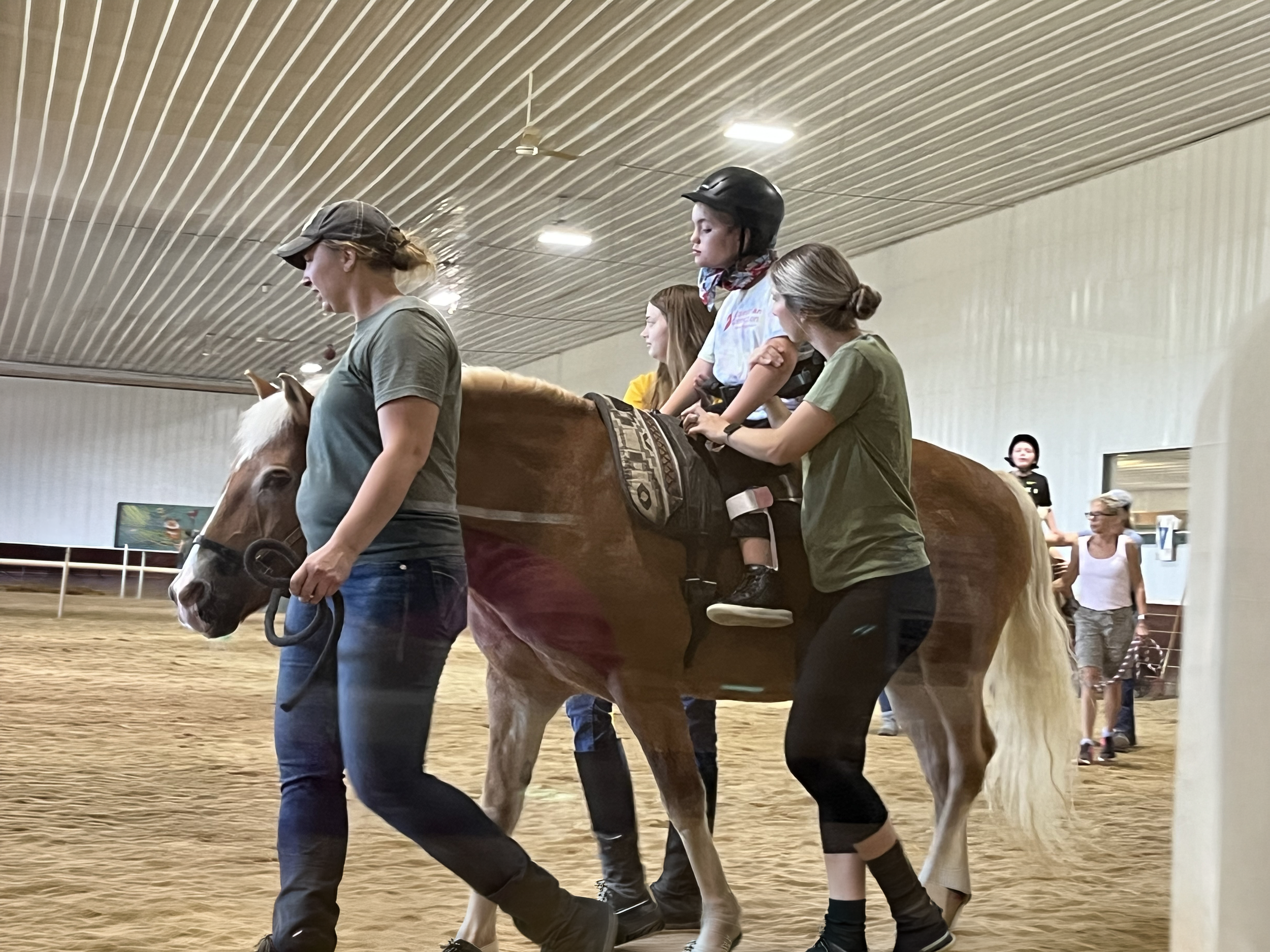 People riding horses inside an indoor riding arena, with some individuals leading and guiding the horses while others ride. The ceiling is metallic with strip lighting.