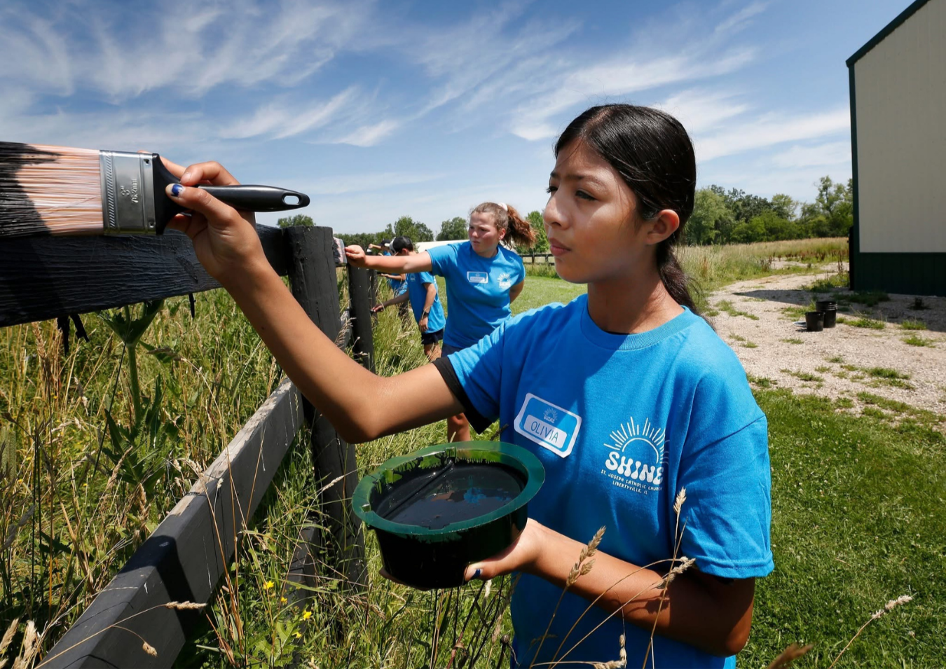 A girl named Olivia painting a black wooden fence blue with a paintbrush, with other children in blue shirts in the background, outdoors on a sunny day.