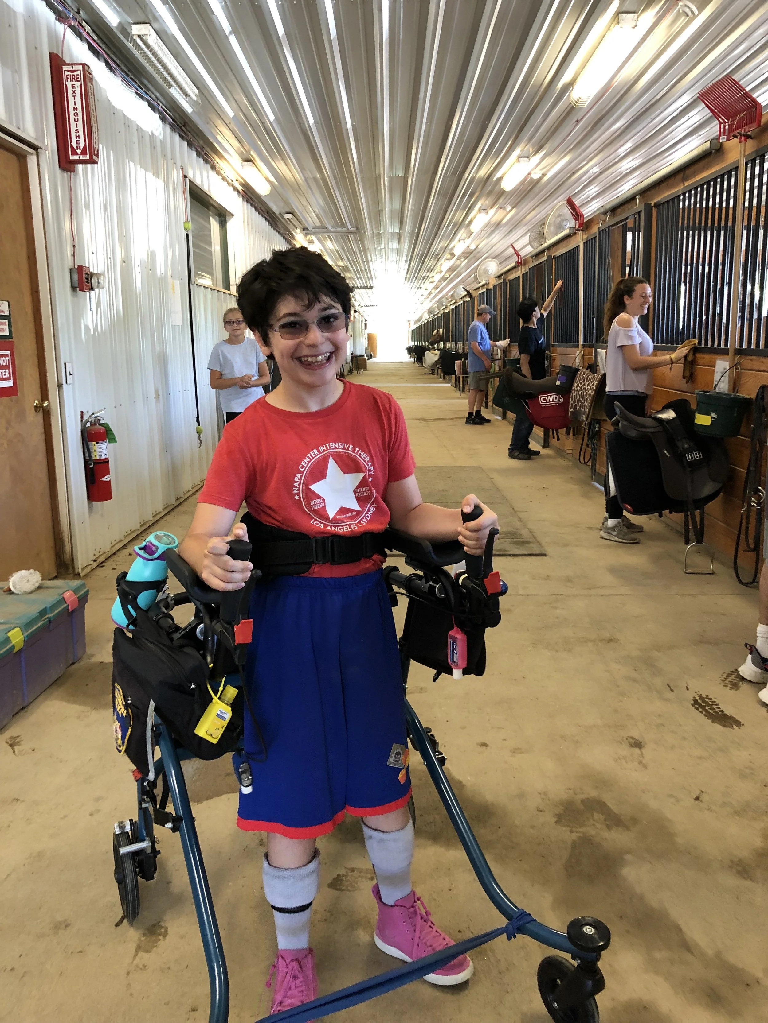 Smiling boy in a red shirt and blue shorts standing with a walker inside a barn or stable, with others in the background.