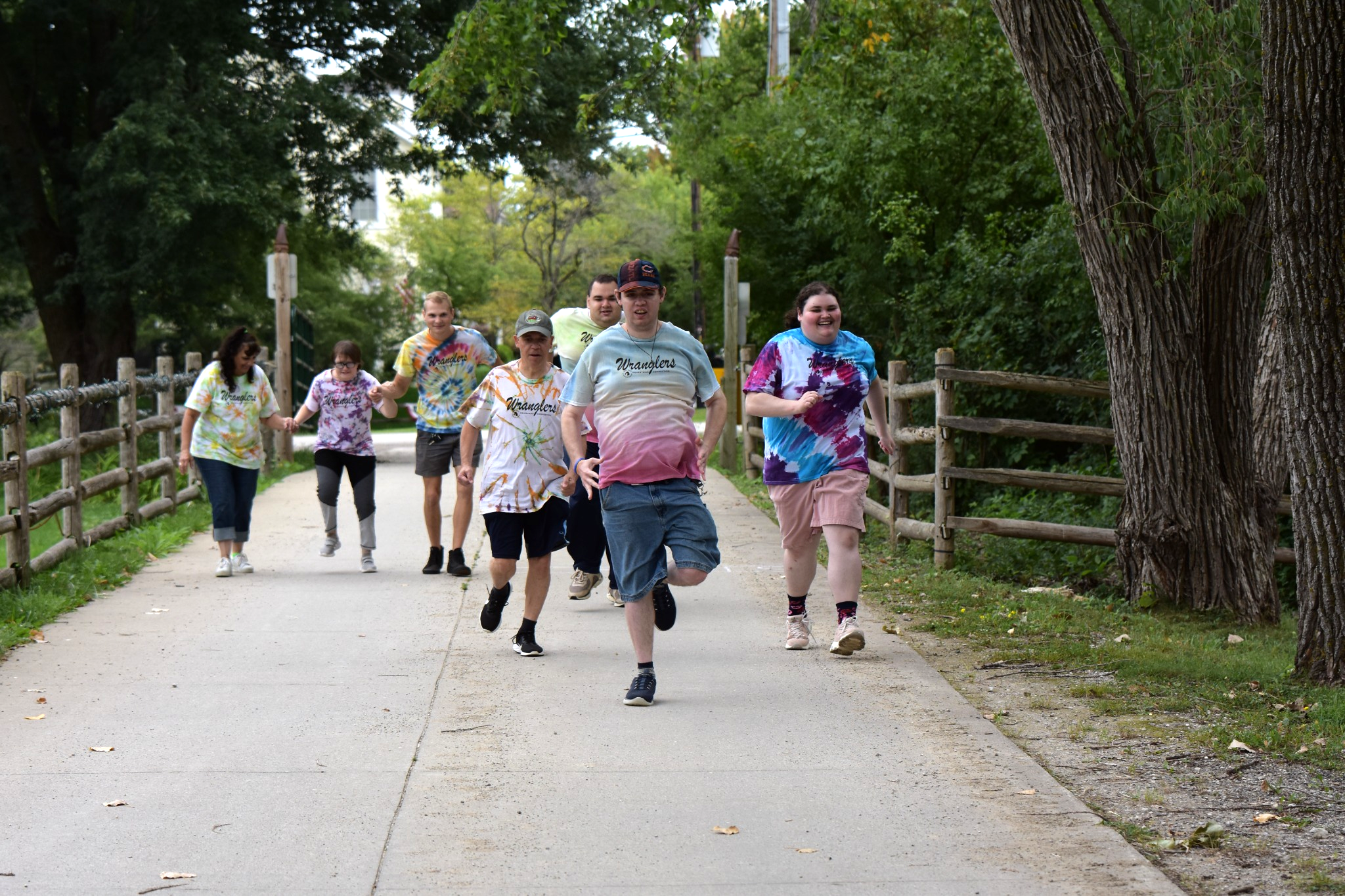 A group of seven people walking and running along a paved path in a park surrounded by trees and greenery, with some wearing colorful tie-dye shirts and smiling.
