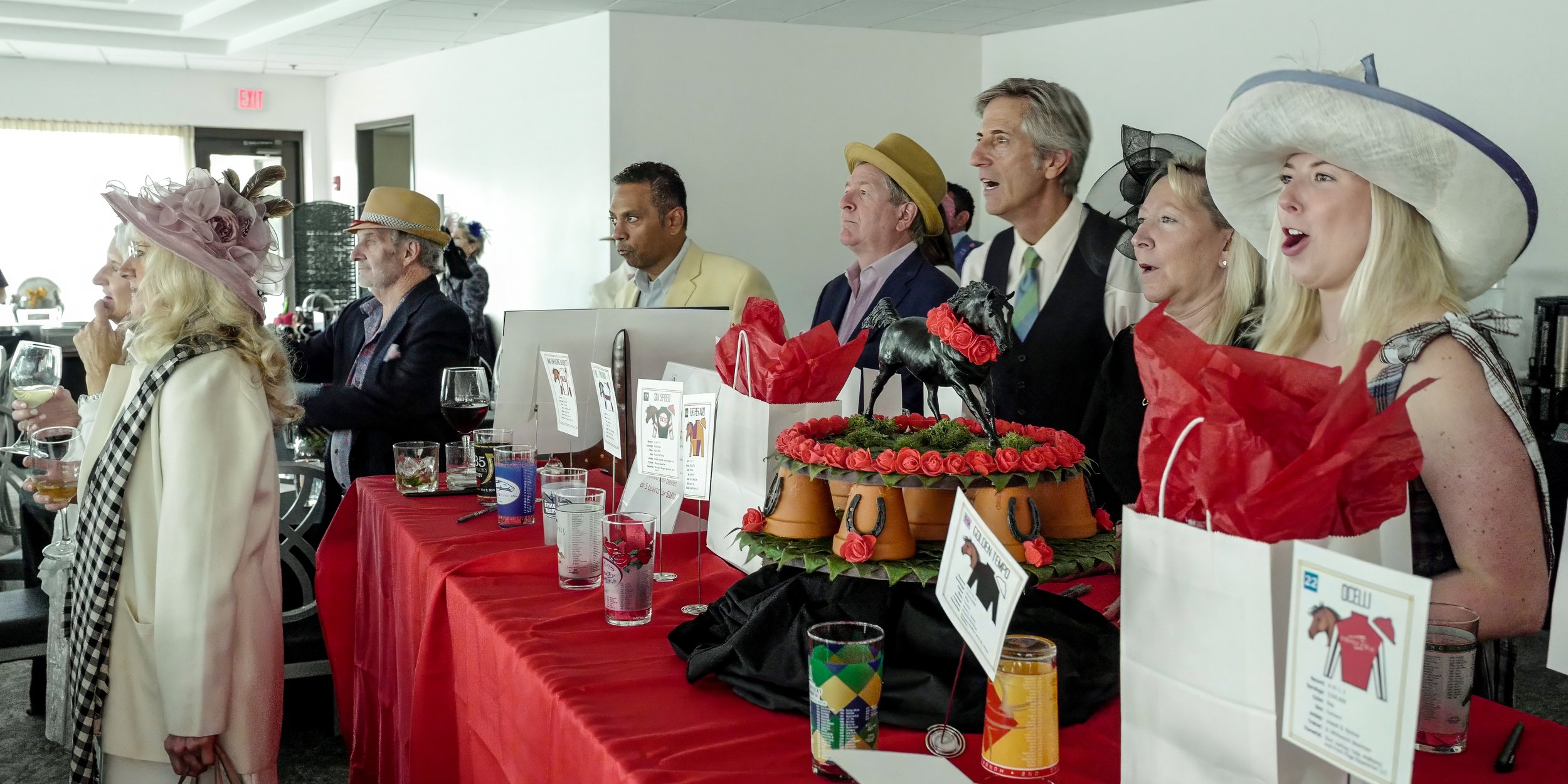 Group of people standing behind a red table with decorative items, some holding drinks, in a bright indoor space.