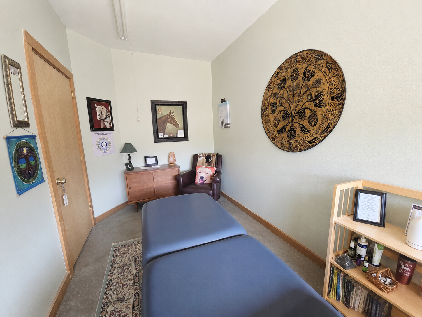 A room with light-colored walls decorated with horse-themed artwork and Celtic wall art. Contains a wooden sideboard with a lamp, framed photo, salt lamp, and a picture of a dog pillow on a leather armchair. A massage table is in the foreground, and a wooden bookshelf with books, skincare products, and decorative items is on the right. The room has a door with a calendar hanging next to it and a small mirror on the wall.