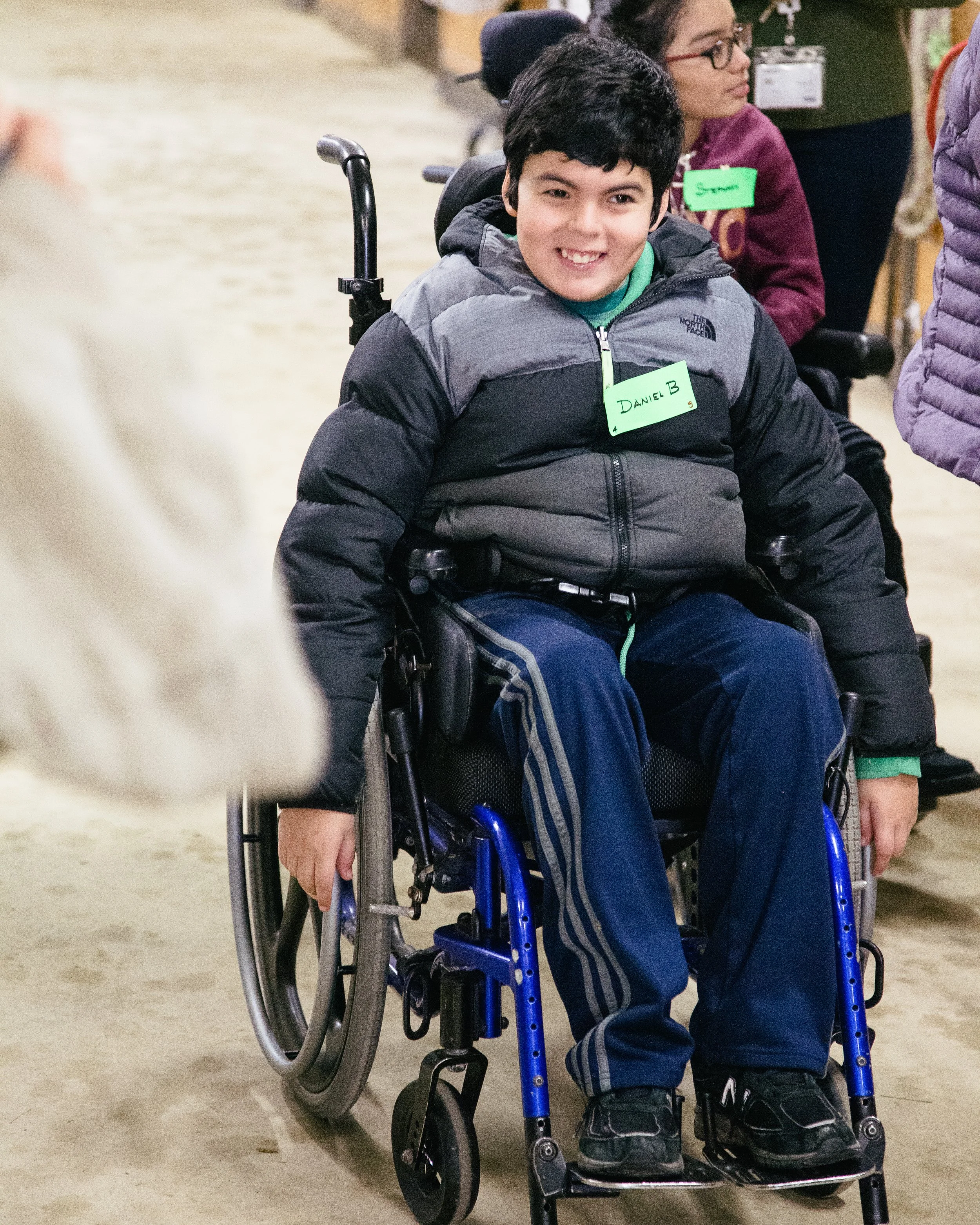 A young person with short black hair smiling while sitting in a wheelchair, wearing a black and gray North Face puffer jacket, dark blue pants, and black shoes. In the background, a woman with glasses and short brown hair is wearing a purple jacket and a green name tag.