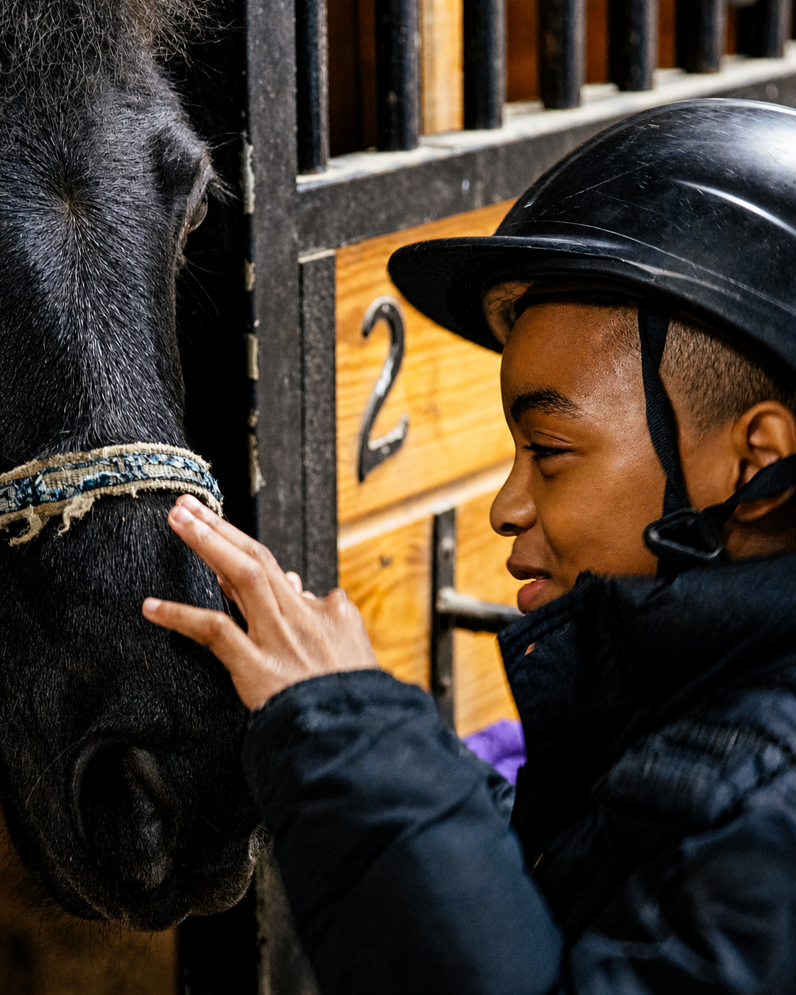 A person with a riding helmet gently touching a horse's face inside a stable.