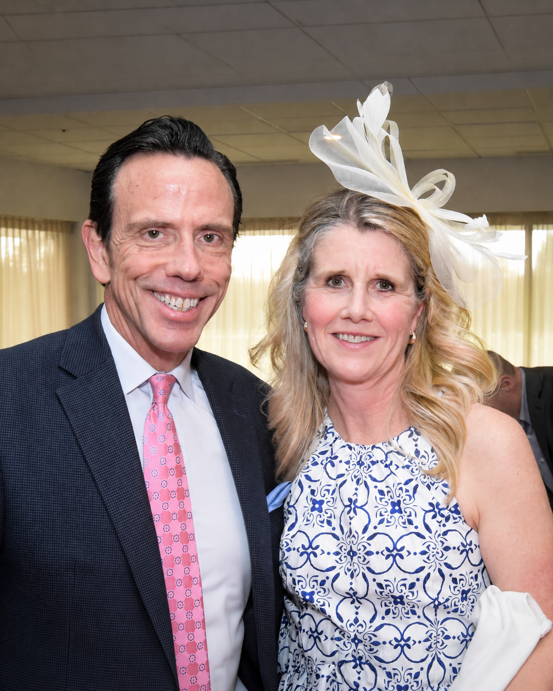 A man in a dark suit and pink tie standing beside a woman in a patterned dress and large white fascinator hat at an indoor event.