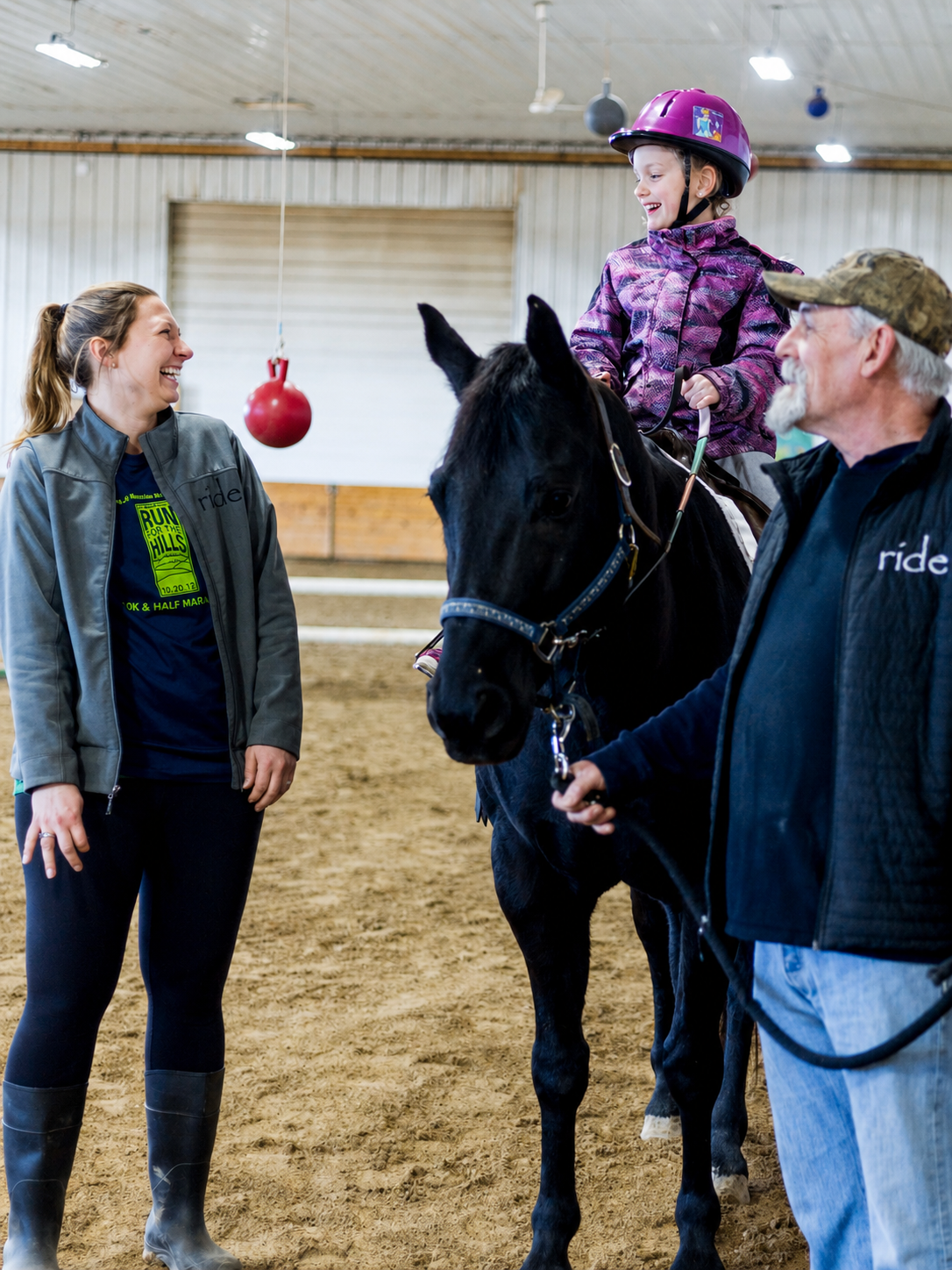 A young girl on a black horse wearing a purple helmet, smiling, with two adults, a woman and a man, in an indoor riding arena. The woman is laughing, and the man is holding the horse's lead. A red ball is hanging from the ceiling.