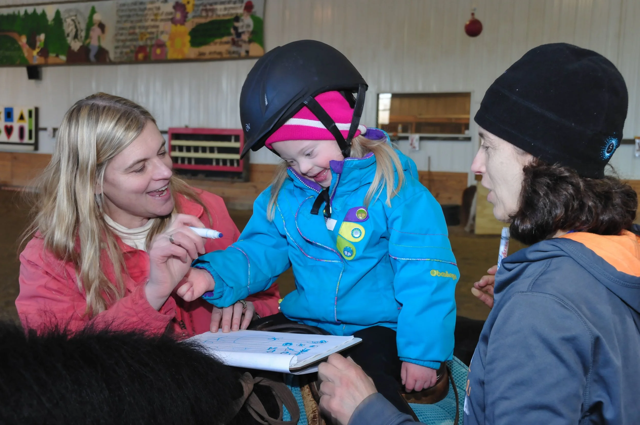 A young girl in winter clothing with a helmet, smiling and interacting with two adults, one woman and one man, in an indoor setting with colorful decorations and a mural in the background.