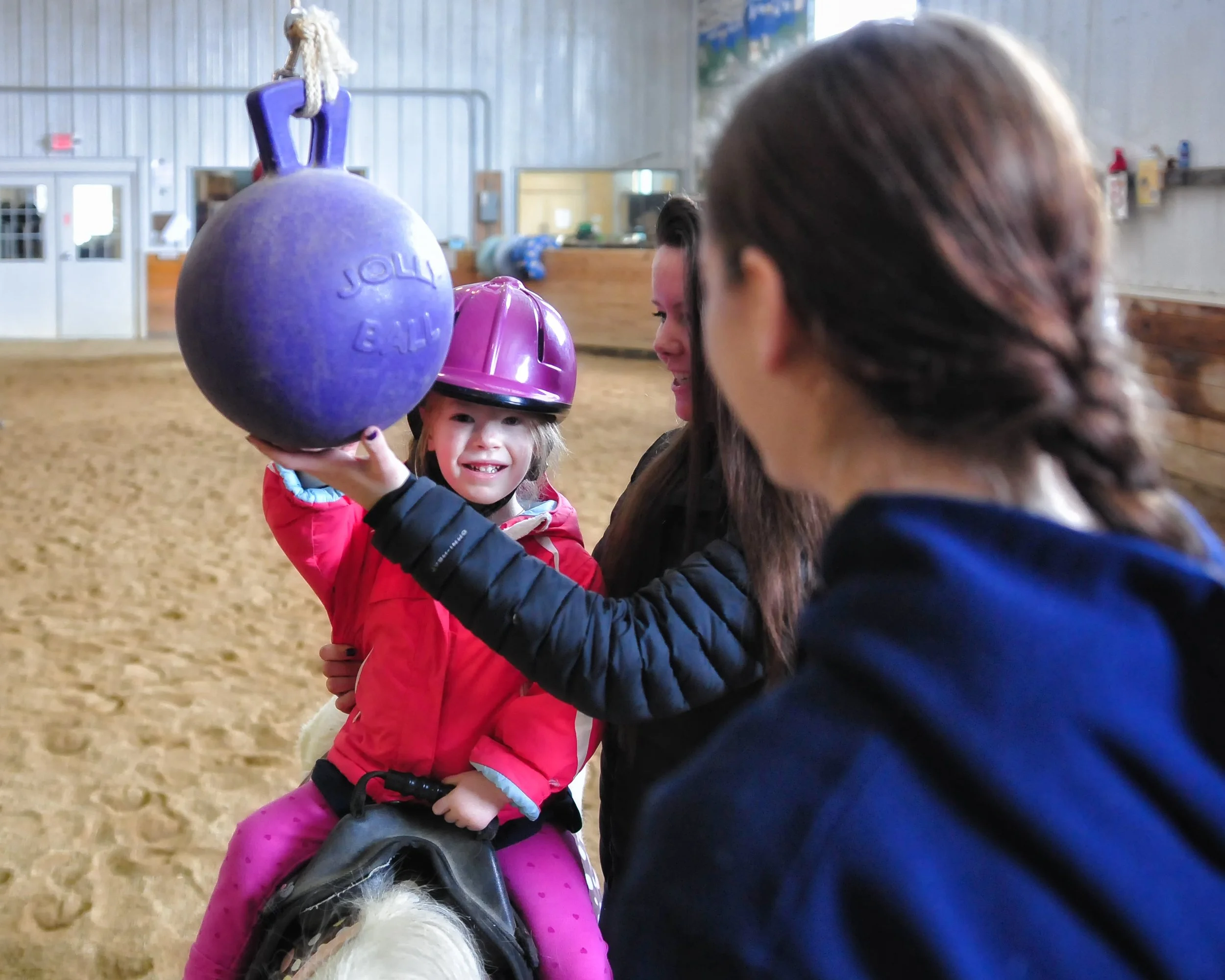 A young girl sitting on a horse with a helmet, smiling, wearing a pink jacket and pink pants, while adults stand nearby inside an indoor riding arena.
