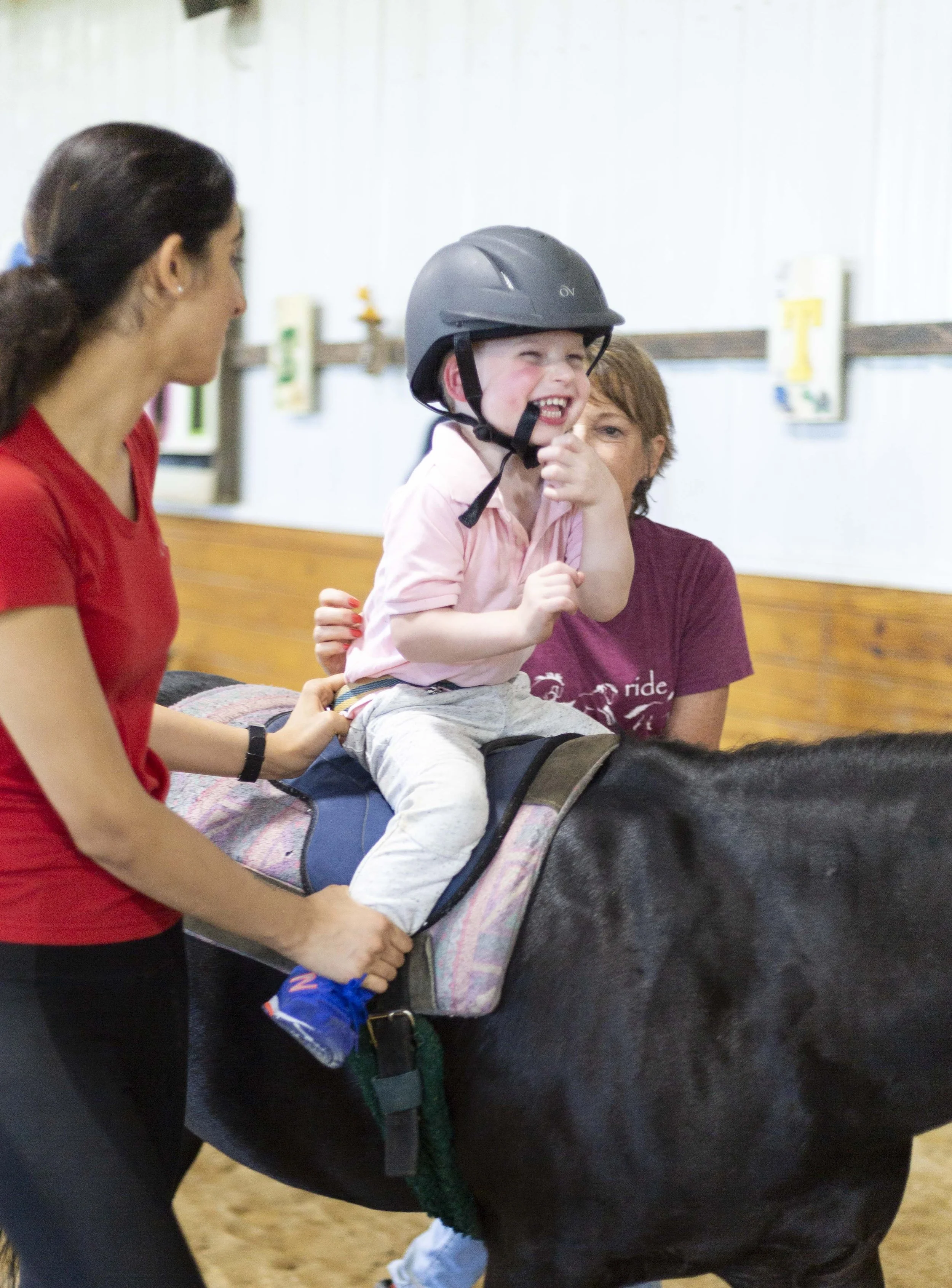 A young child wearing a helmet sits on a horse, smiling joyfully, while being assisted by adults.