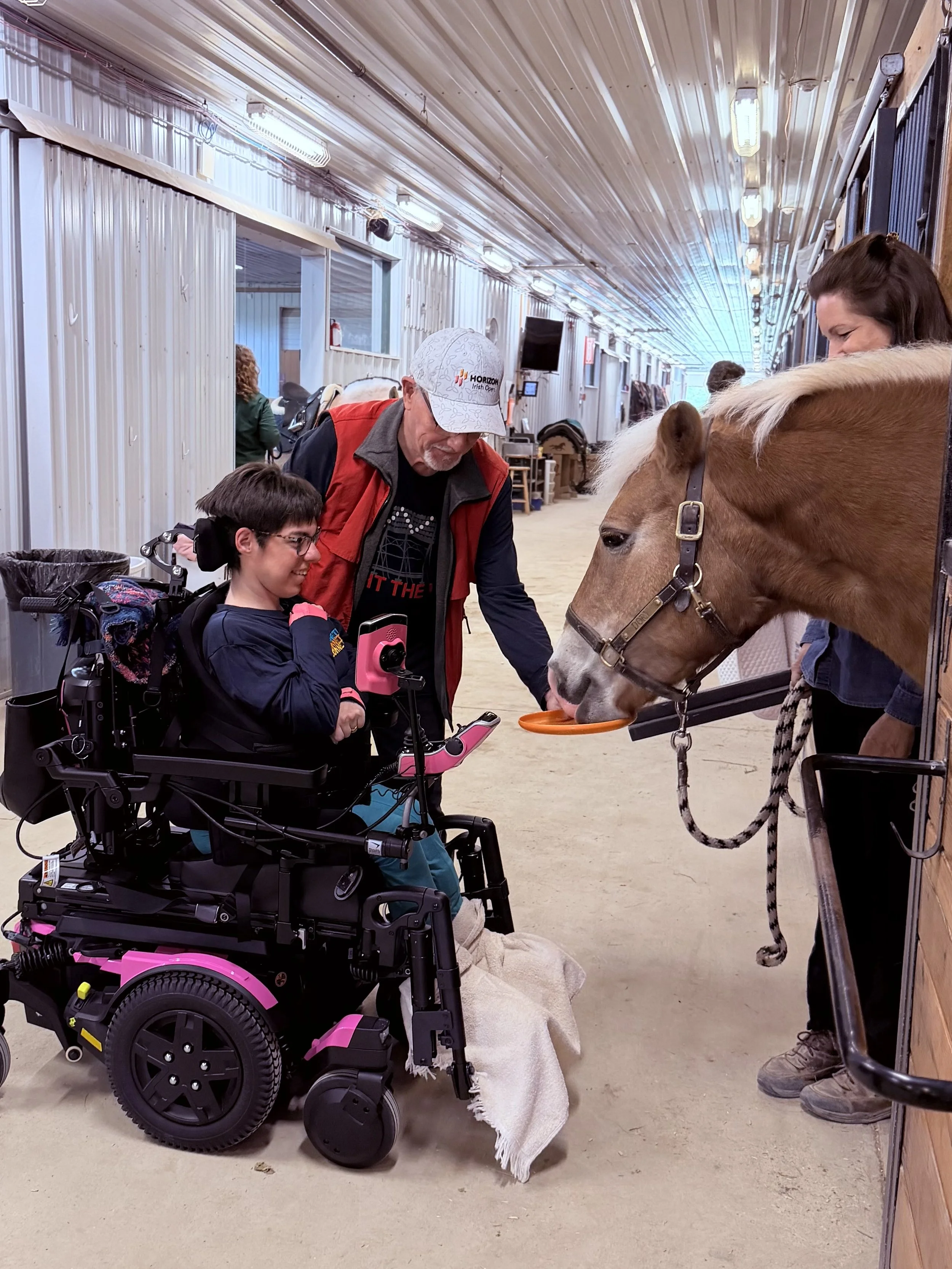 A person in a wheelchair and a person with a horse, inside a barn. The horse is eating from a bowl, and the person in the wheelchair is looking at the horse. Two other people are visible in the background.