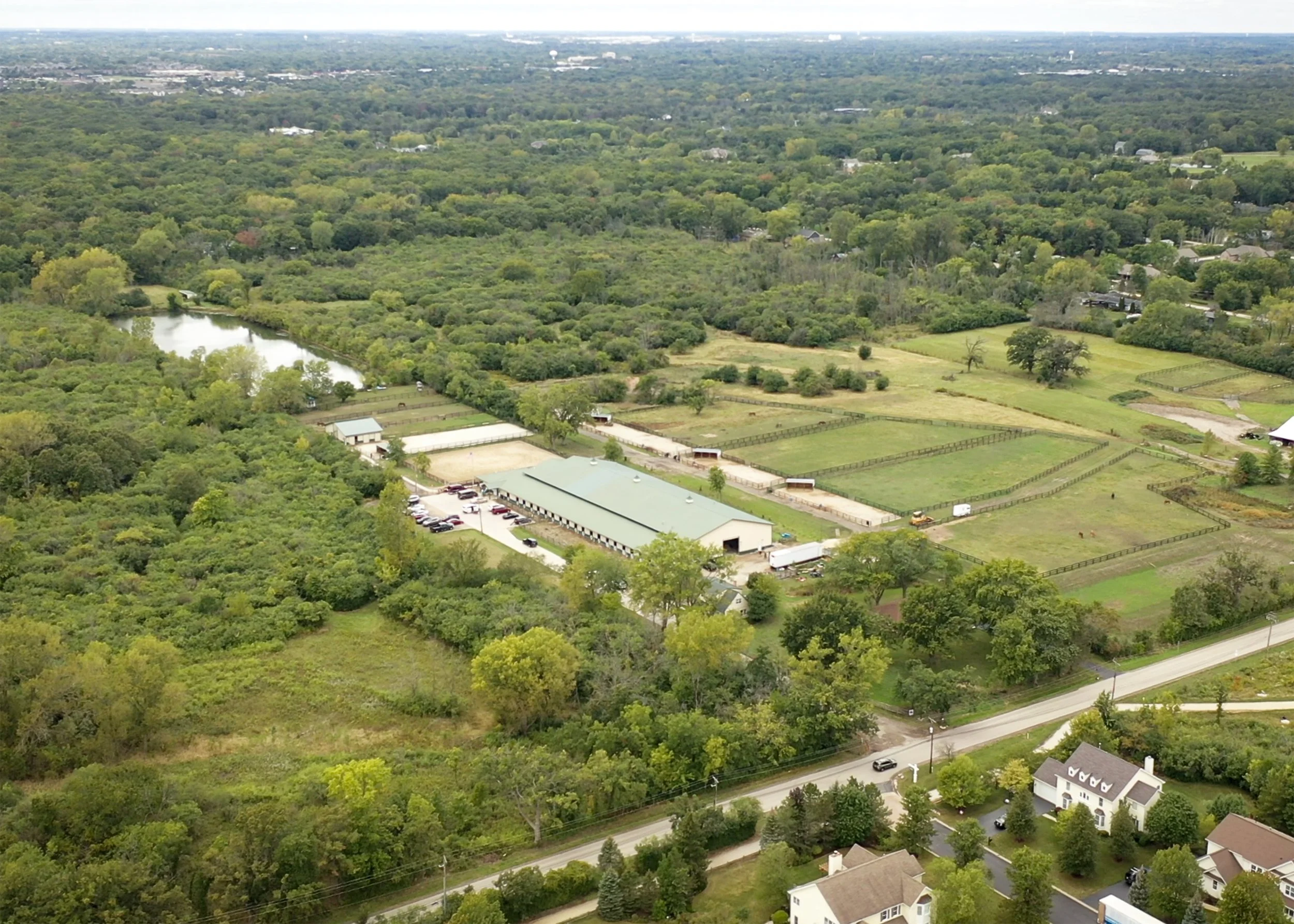 Aerial view of a rural area with dense green trees, a pond, open fields, and residential houses under a cloudy sky.