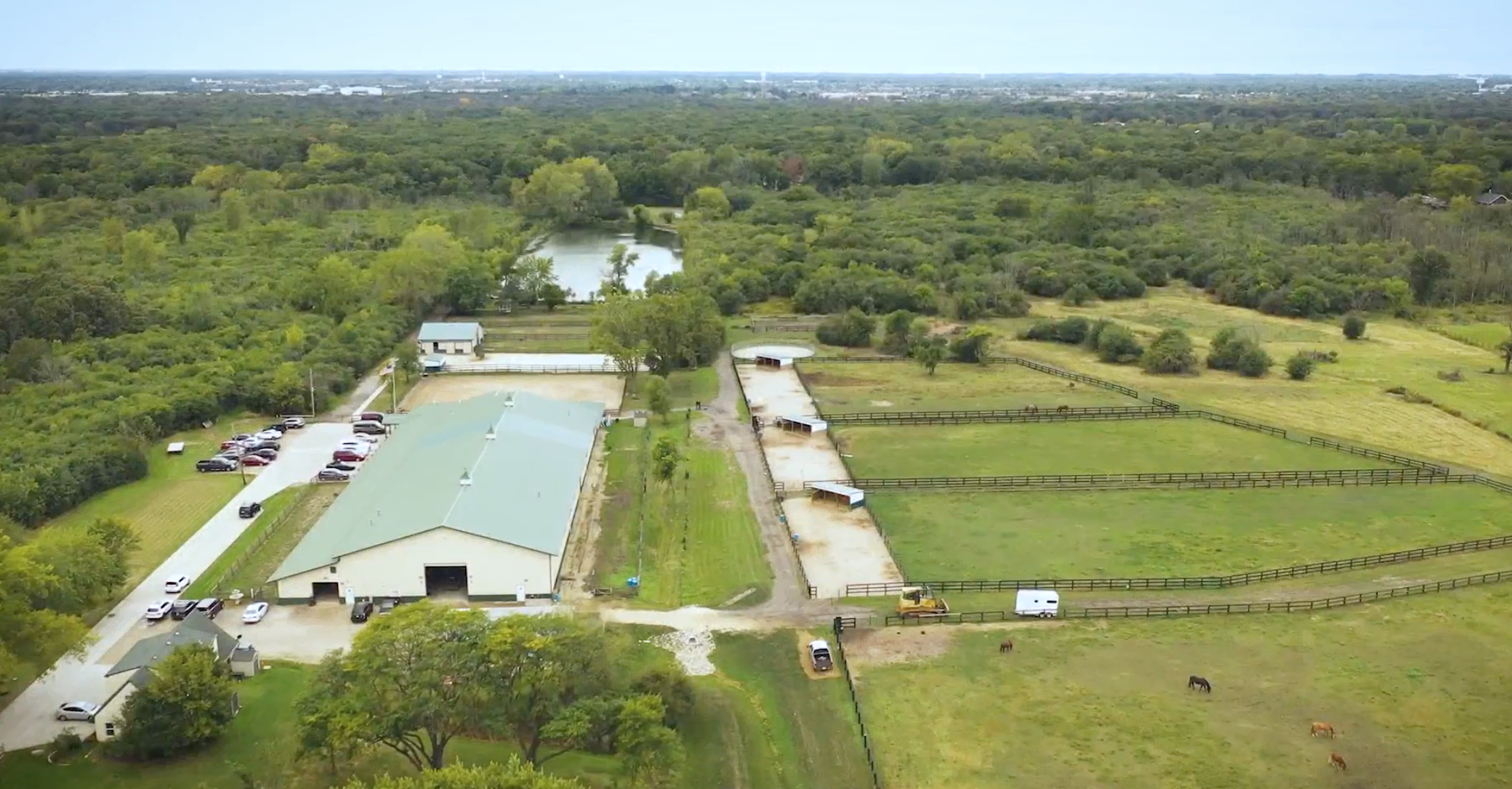 An aerial view of a farm or equestrian facility with covered riding arenas, fenced pastures, a pond, buildings, and parking lot, surrounded by lush green trees and open fields.