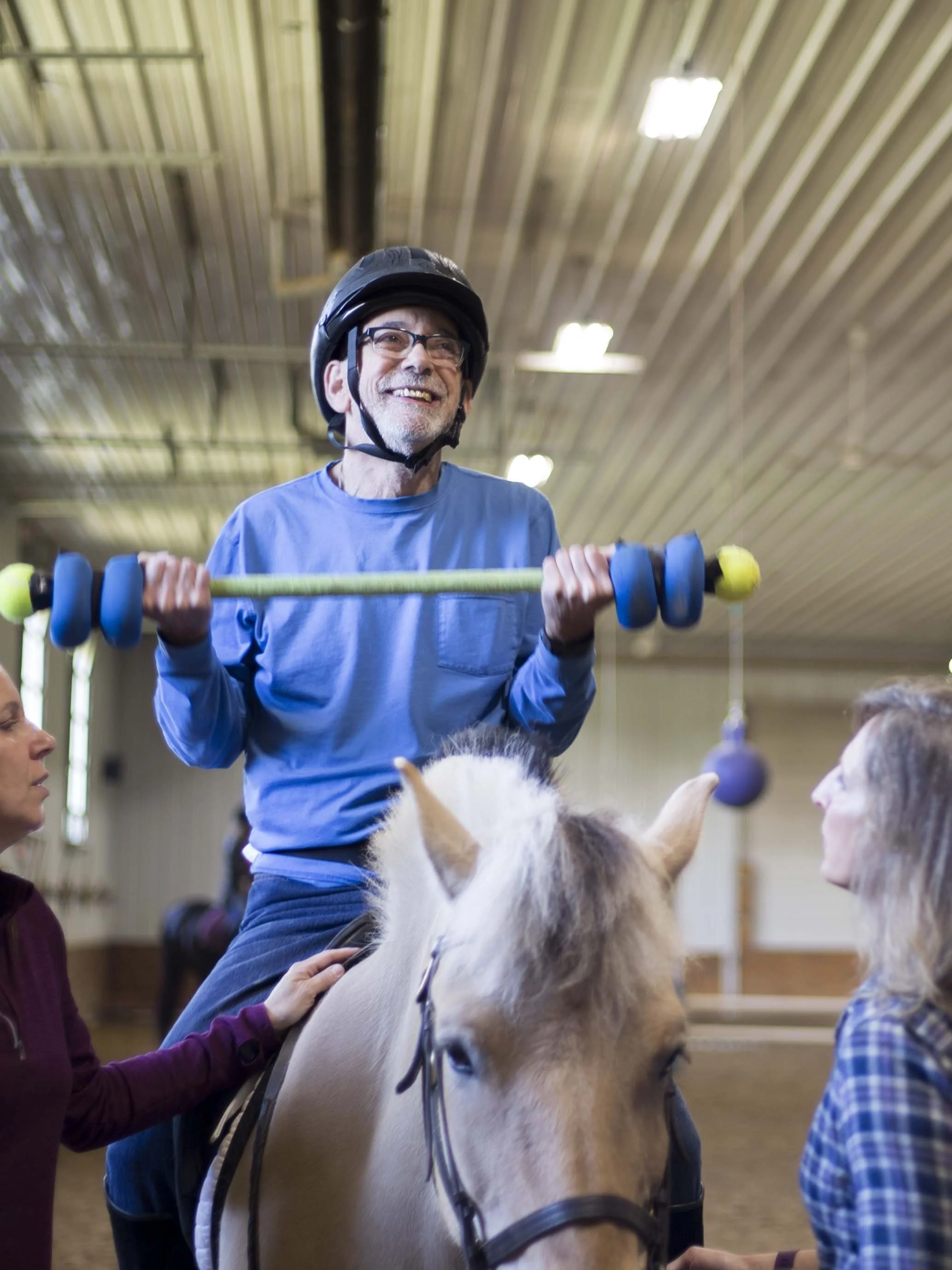 An elderly man wearing a helmet is riding a horse indoors, with a group of people around him, and a woman is holding the horse's reins.