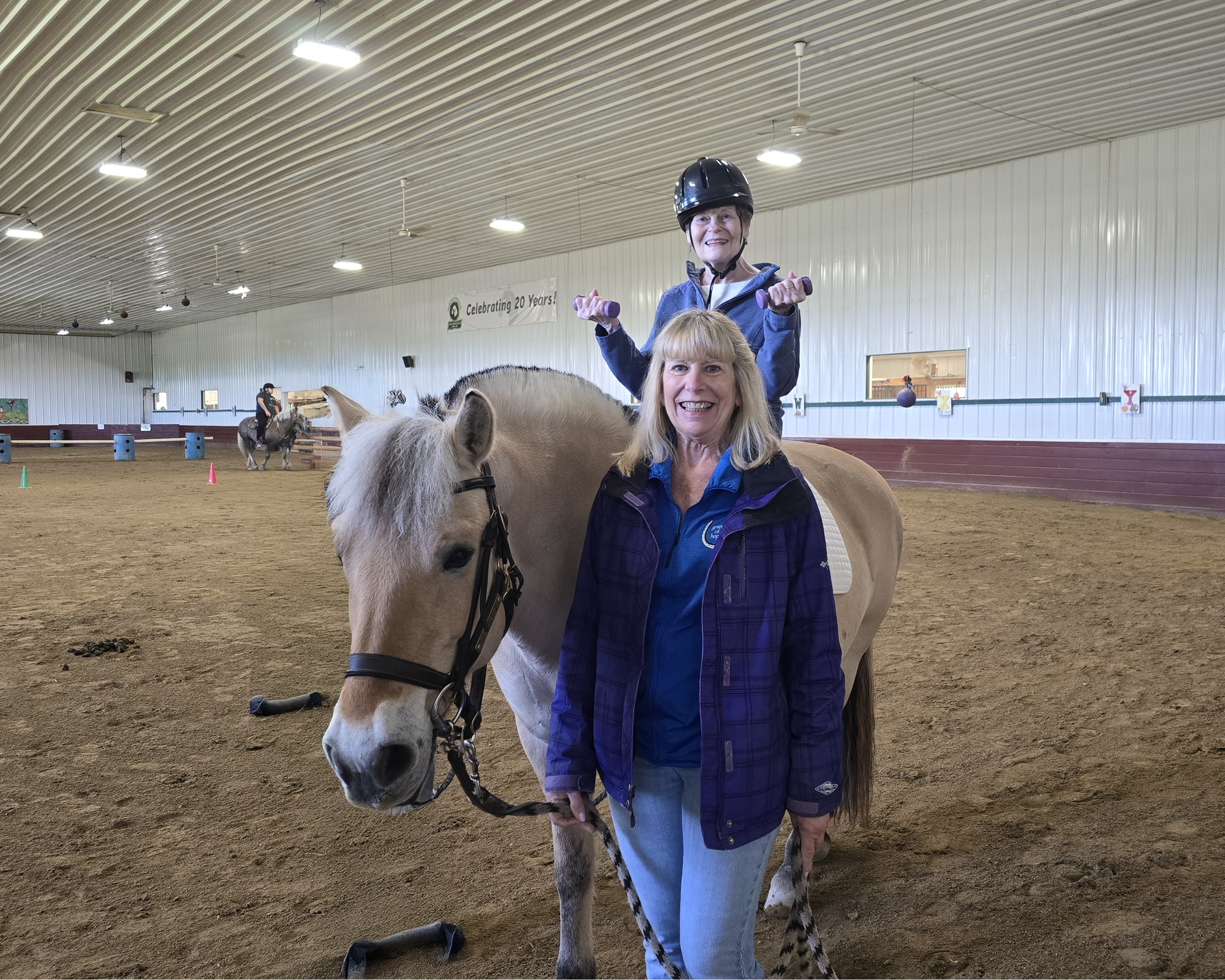 Two smiling women, one older and one younger, posing with a small pinto horse inside an indoor riding arena. The older woman is holding the horse's bridle while the younger woman stands beside it. The younger woman is sitting on the horse's back, wearing a helmet. There are colorful cones on the ground and a banner on the wall that celebrates 20 years.