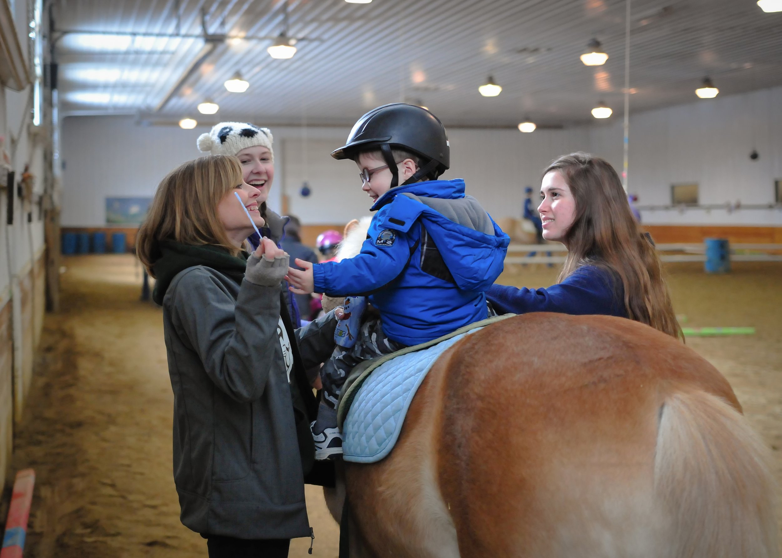 A woman is sitting on a horse while children stand nearby in an indoor riding arena. One child is wearing a helmet and smiling at the woman, and two other children are watching.