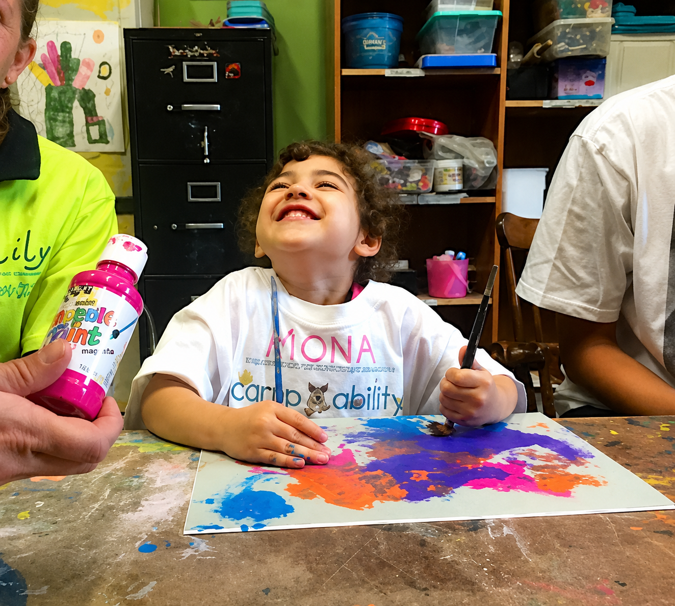 A young girl with curly hair smiling happily at an art table with colorful paint on paper in front of her. An adult holding a pink bottle of paint is partially visible on the left, and child-sized chairs and shelves with art supplies are in the background.