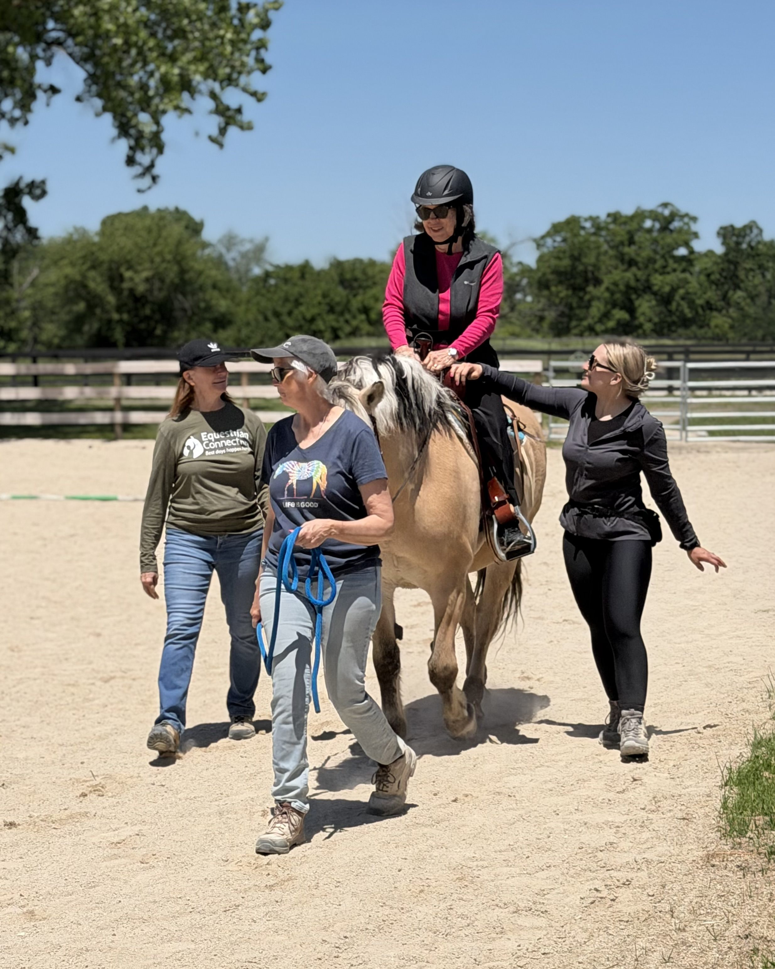 A group of women, some wearing sunglasses and hats, standing around and guiding a woman riding a small horse or pony in an outdoor riding arena. The woman on the horse is wearing a helmet and a pink and black vest, smiling, with trees and a blue sky in the background.
