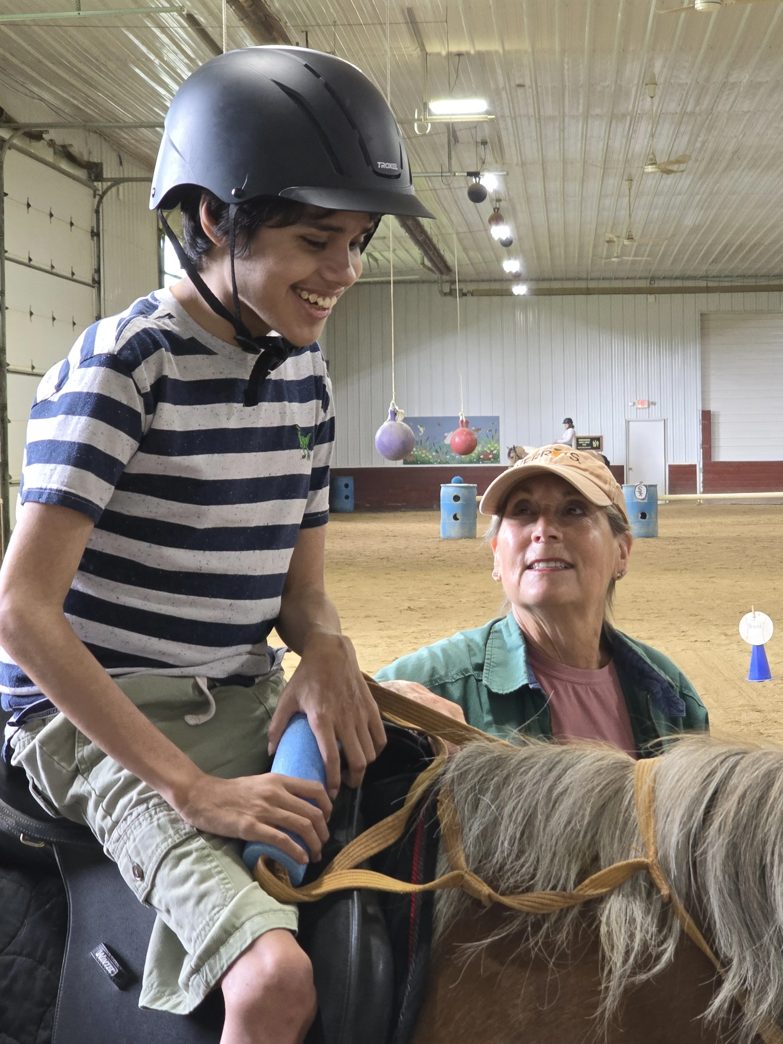 A young person wearing a black helmet, sitting on a horse, and smiling while talking to an older woman in a plaid shirt inside an indoor equestrian riding arena.