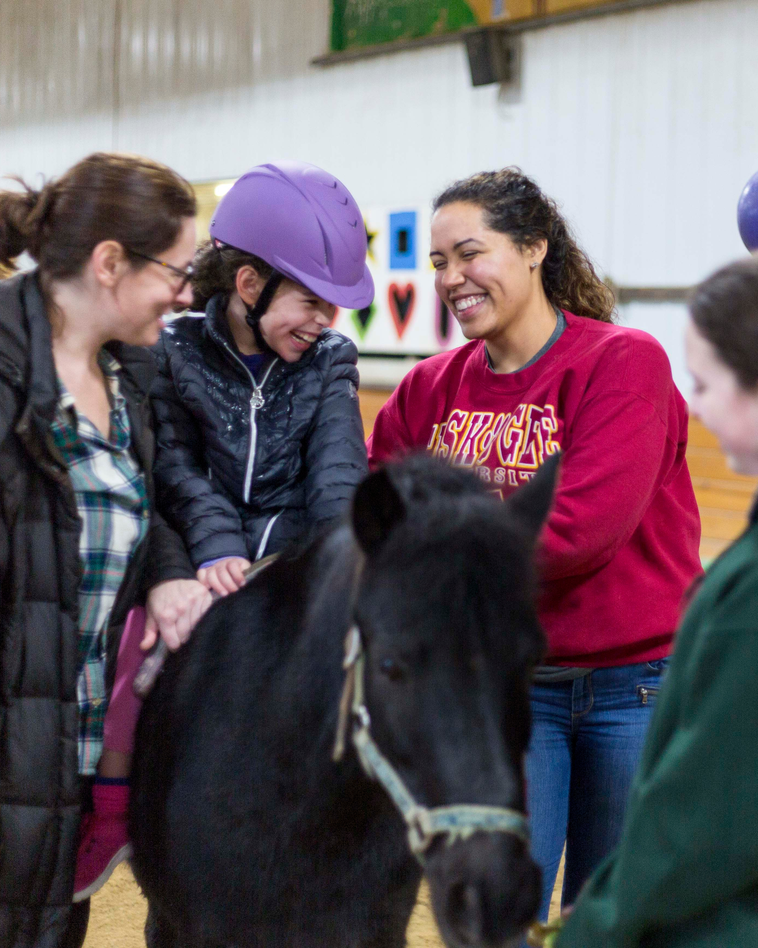 Group of people, including children, gathered indoors, smiling and engaging in conversation. One person is wearing a purple helmet and a puffy jacket, while others wear casual clothing. The background has colorful decorations and a white fence.