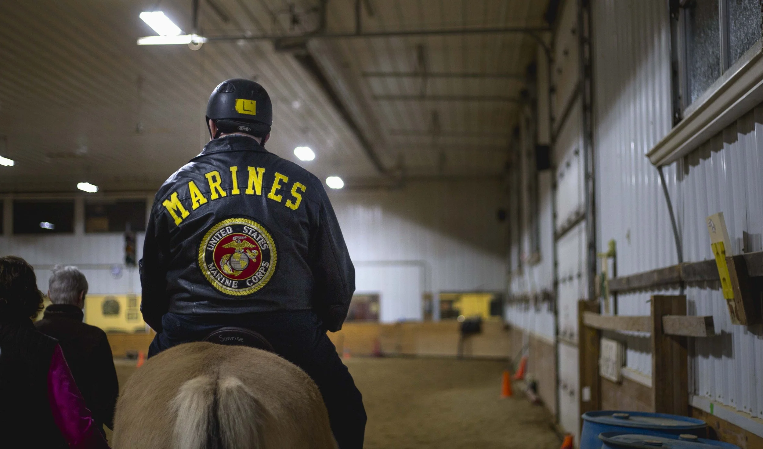 A person wearing a black jacket with yellow text 'MARINES' and the Marine Corps emblem on the back, and a helmet with a yellow badge, is riding a horse inside an indoor riding arena, with spectators sitting nearby and orange cones on the ground.