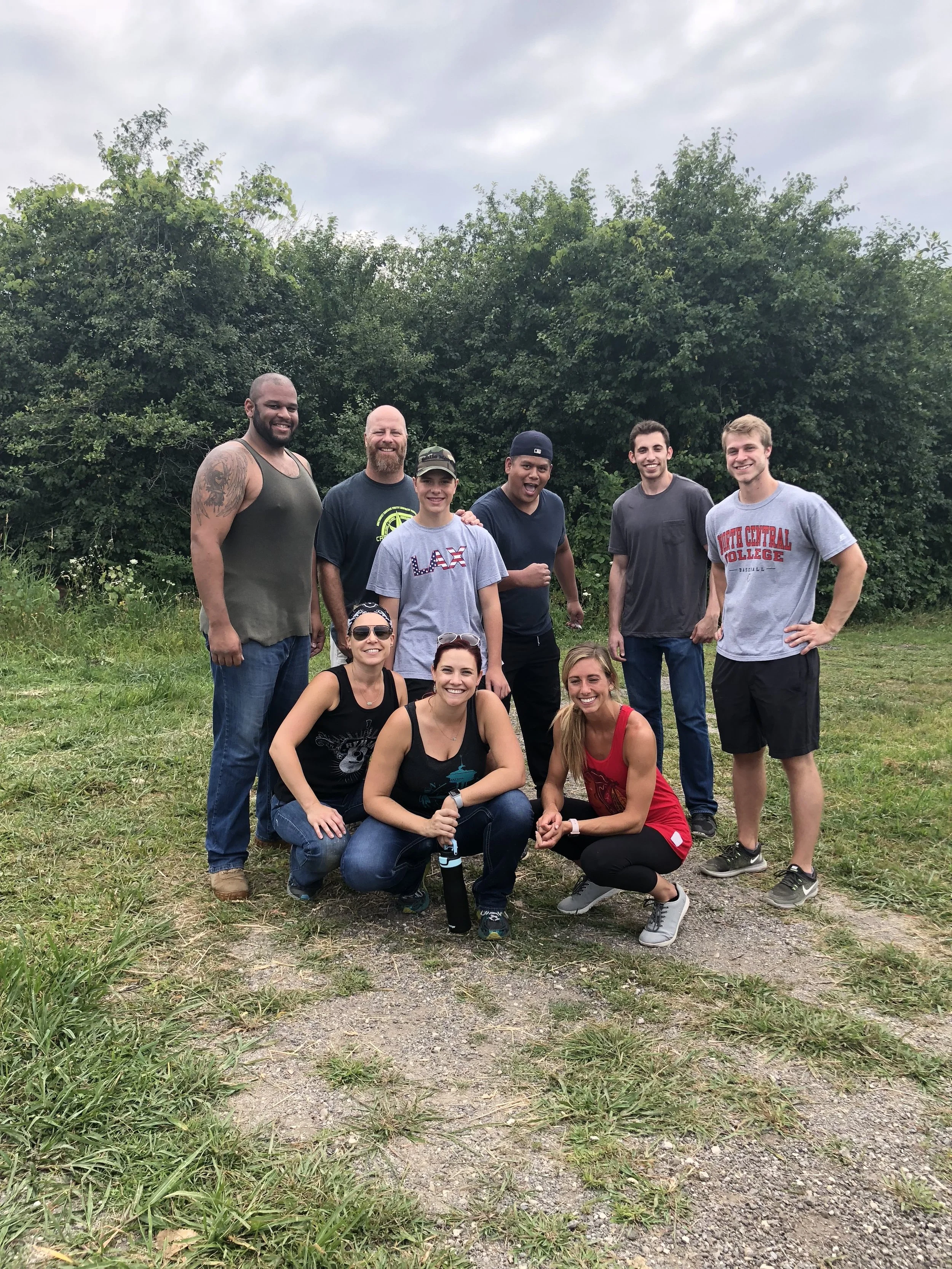 A group of ten smiling young adults posing outdoors near a grassy area with trees in the background, wearing casual clothes and sunglasses.