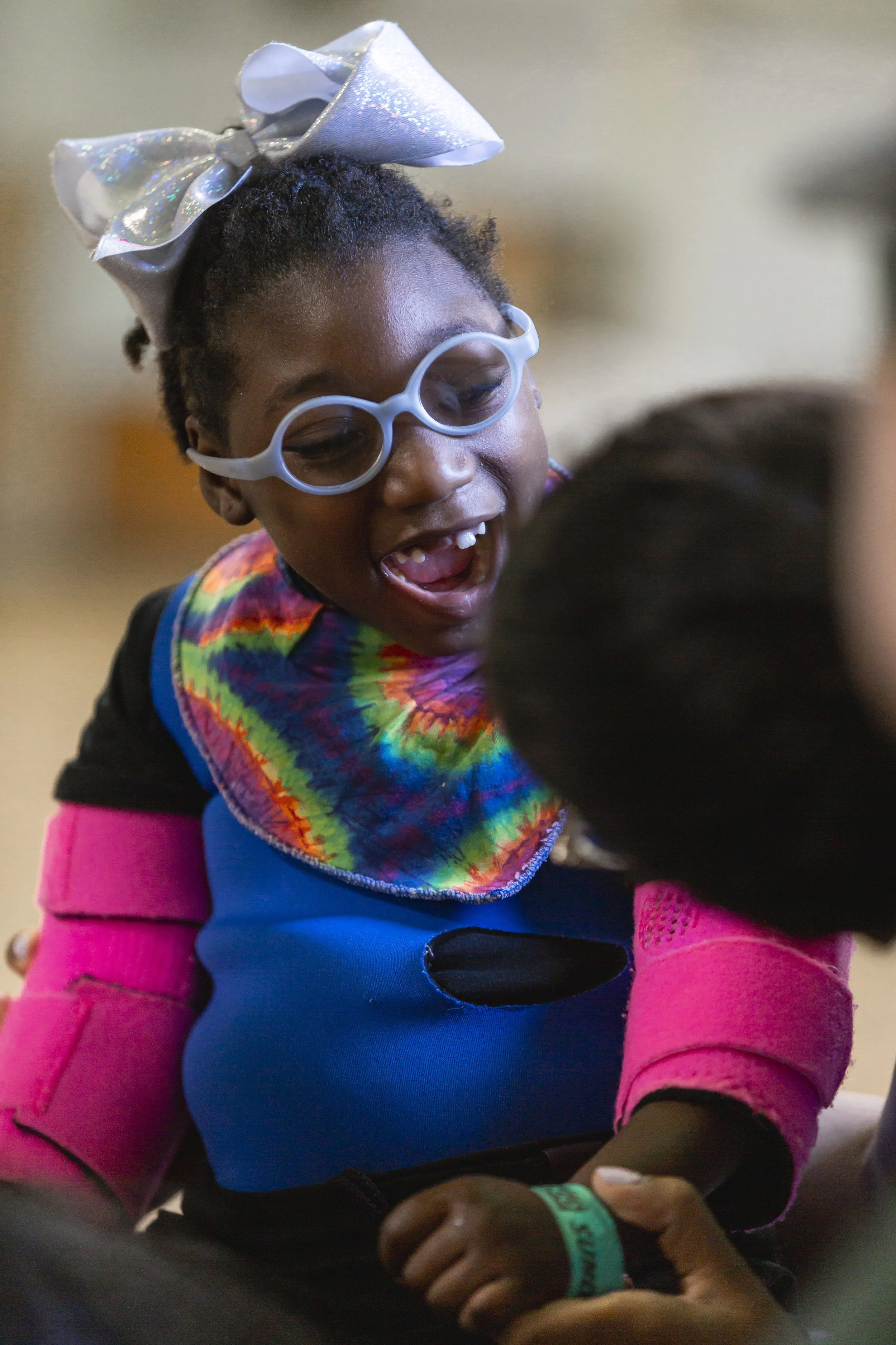 A young girl with curly hair wearing a large shiny silver bow, rainbow tie-dye scarf, pink arm warmers, and light purple glasses, smiling and showing off her missing front teeth.