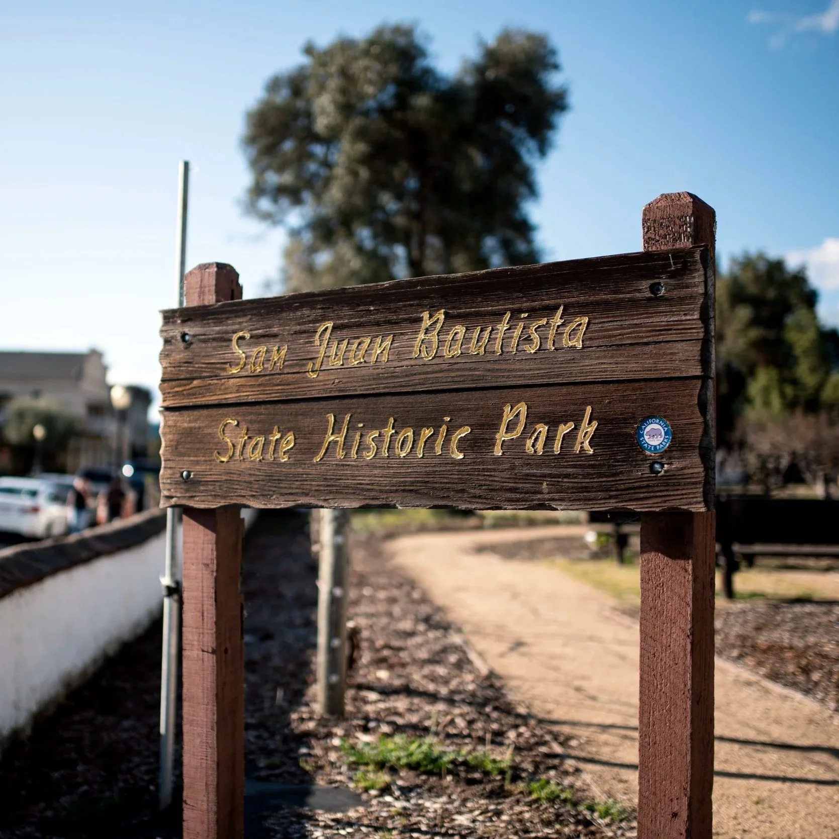 A wooden sign at San Juan Bautista State Historic Park with gold lettering, mounted on two wood posts, in a sunny outdoor setting with trees and the park entrance beyond.