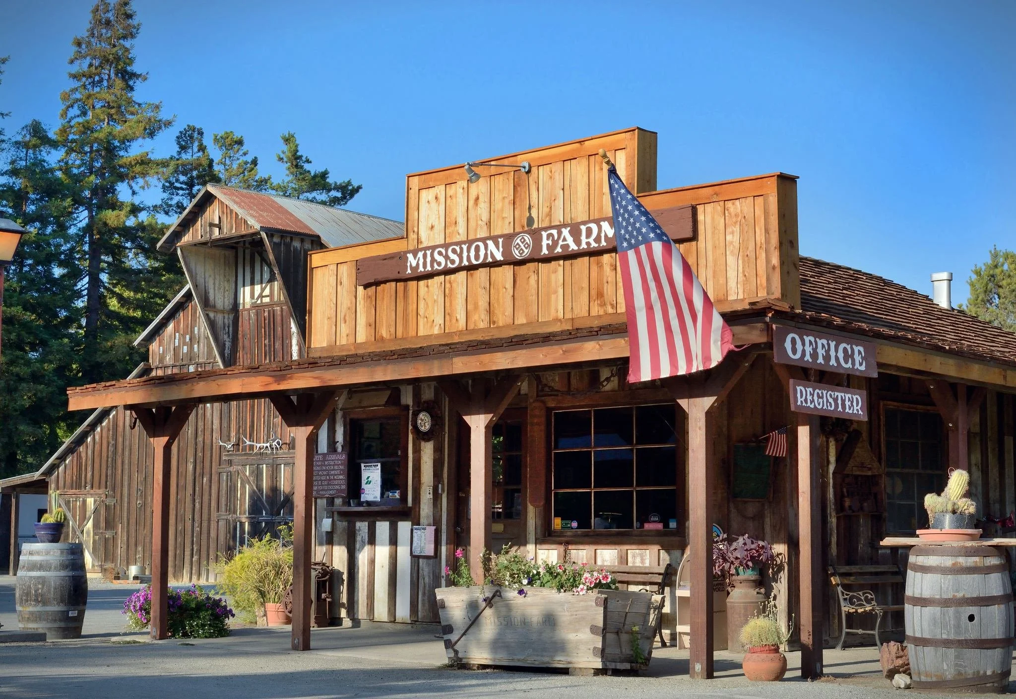 Front view of a rustic wooden building with a sign that reads 'Mission Farm,' decorated with American flags and surrounded by potted plants and barrels, under clear blue sky.