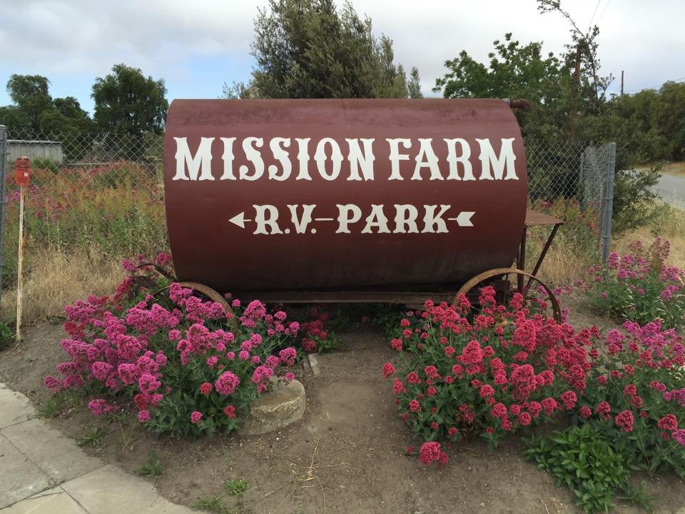 A large, rusty, barrel-shaped sign with white text reading 'MISSION FARM' and 'R.V. PARK' with arrows pointing left and right, respectively. The sign is surrounded by pink flowering plants and located near a chain-link fence and a road in a rural area.