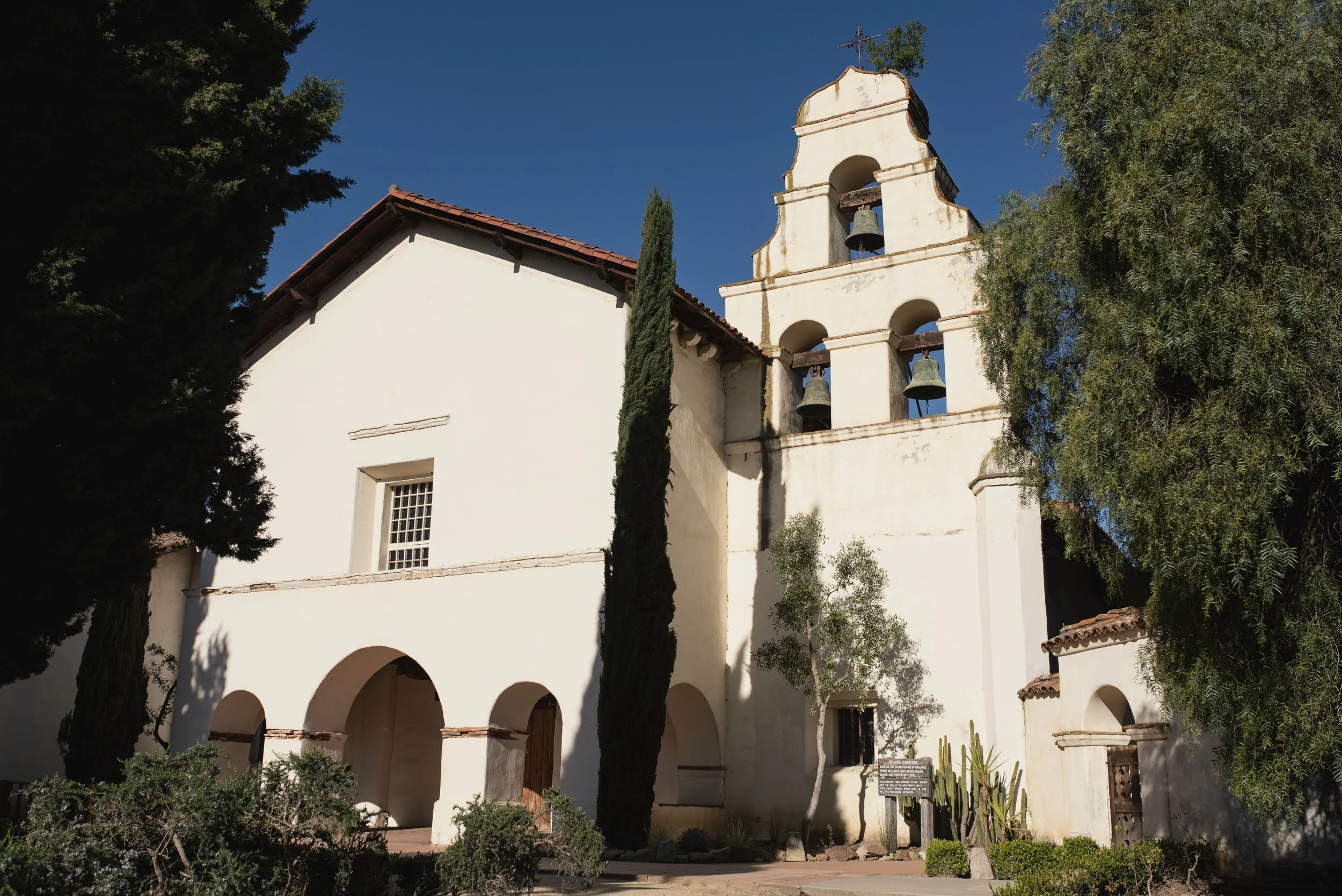 An old white church with a bell tower surrounded by trees and greenery against a bright blue sky.