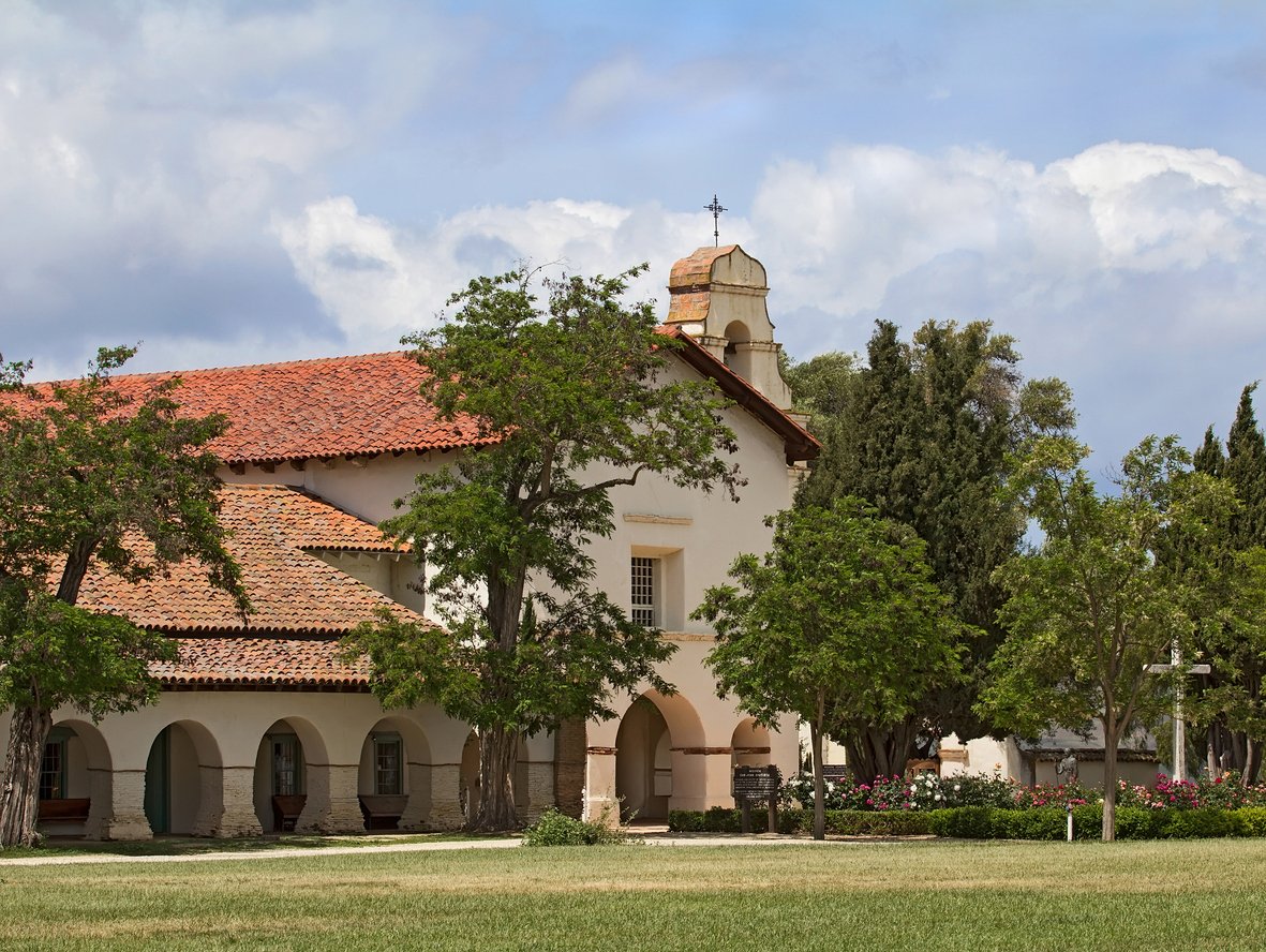 A historic Spanish-style building with white stucco walls, a red tile roof, and arched doorways, surrounded by green trees and a grassy lawn under a partly cloudy sky.