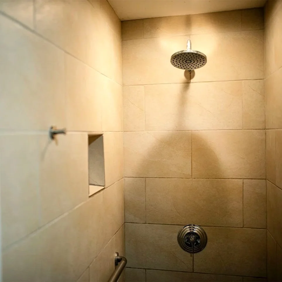 Shower with beige tiles, round showerhead at the top, and a control knob at the bottom, with a partially visible metal grab bar on the left side.