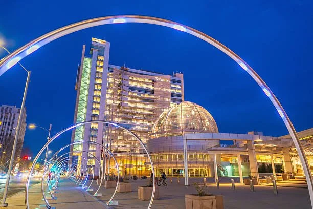 Nighttime cityscape with illuminated modern building featuring a glass dome and a series of decorative arches along a walkway.