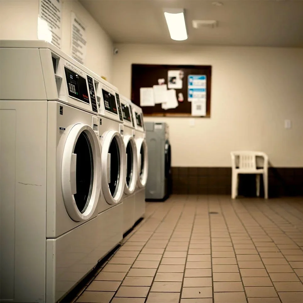 A row of white commercial washing machines in a laundry room with a bulletin board and a white plastic chair in the background.