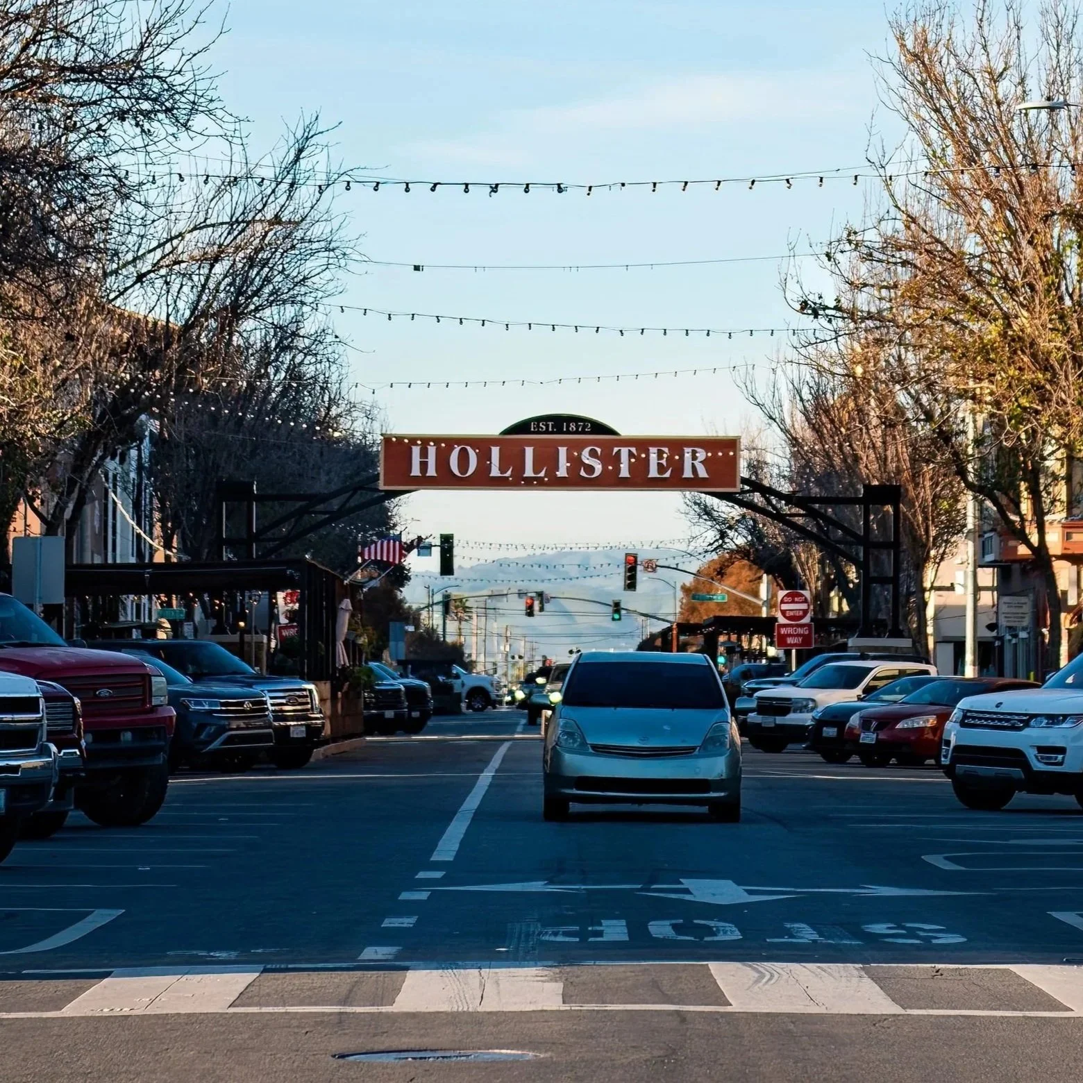 Street view of downtown Hollister with a sign hanging above the road, parking lot with cars, and string lights hanging overhead. Trees line the street, and traffic lights are visible.
