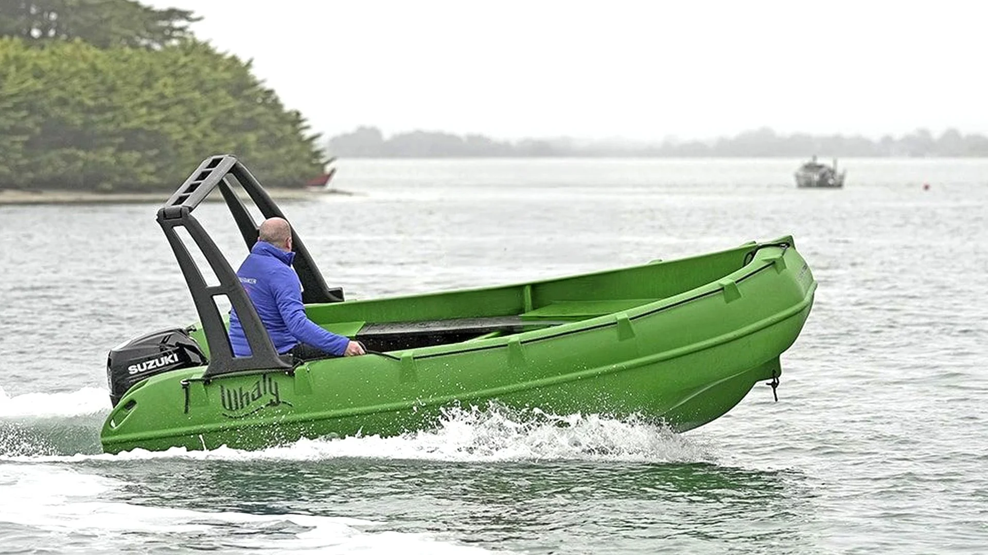 A person in a blue jacket riding a green Whaly boat on a body of water with other boats and trees in the background.
